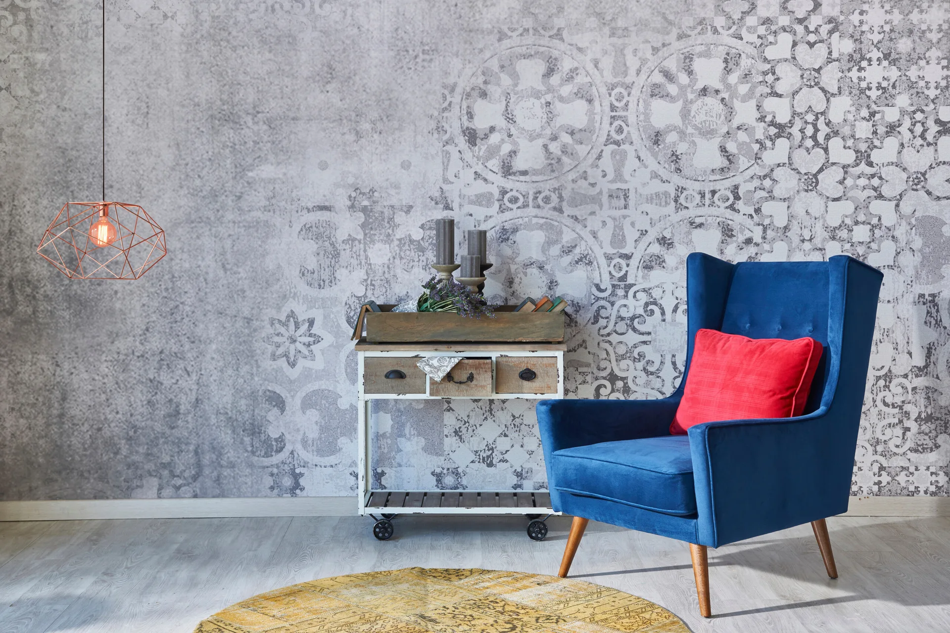 A blue velvet wingback armchair with a red pillow sits on a gray floor next to a wheeled wooden side table against gray patterned wallpaper.