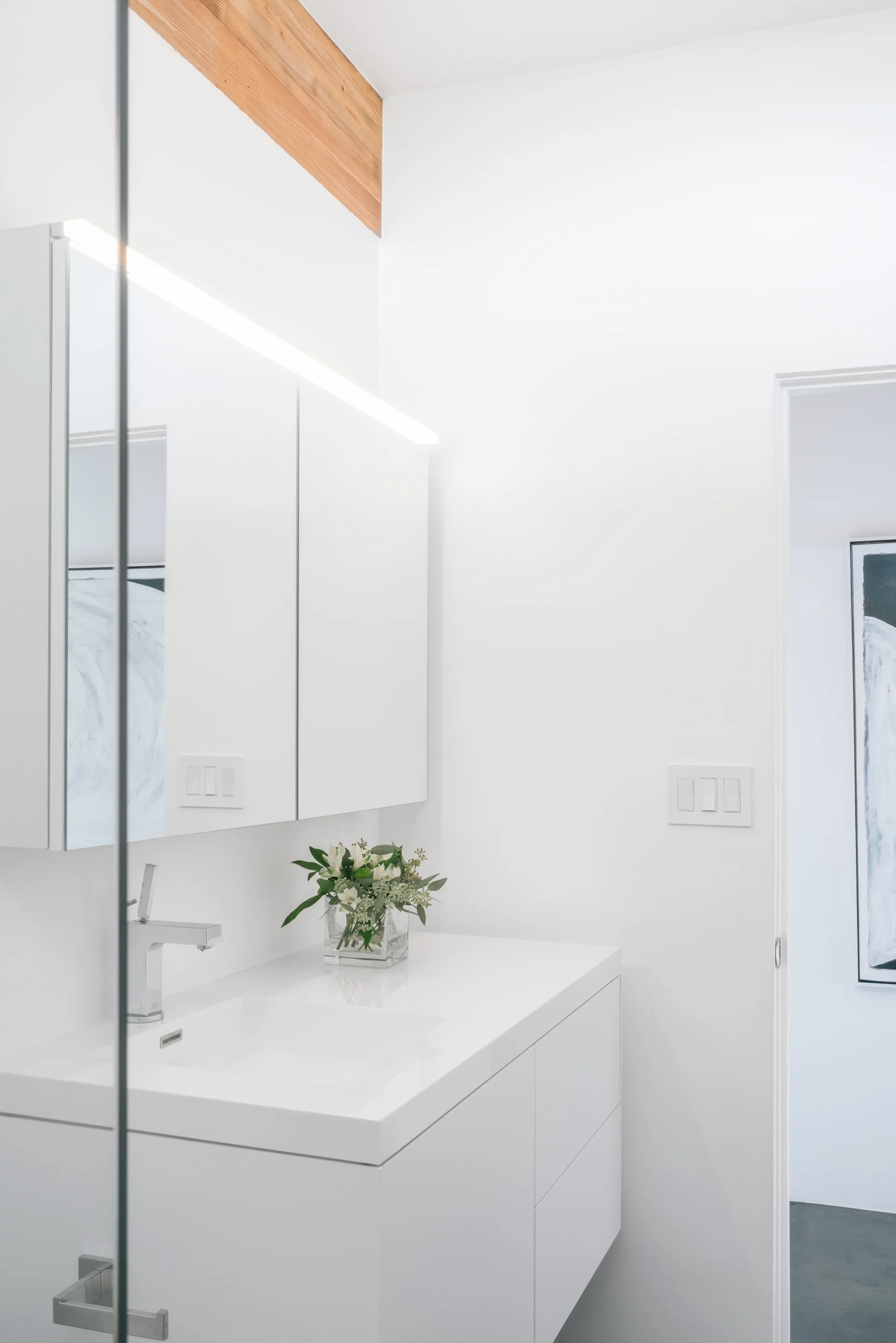 A modern white bathroom features a floating vanity, a mirrored medicine cabinet with integrated lighting, and a natural wood ceiling beam.