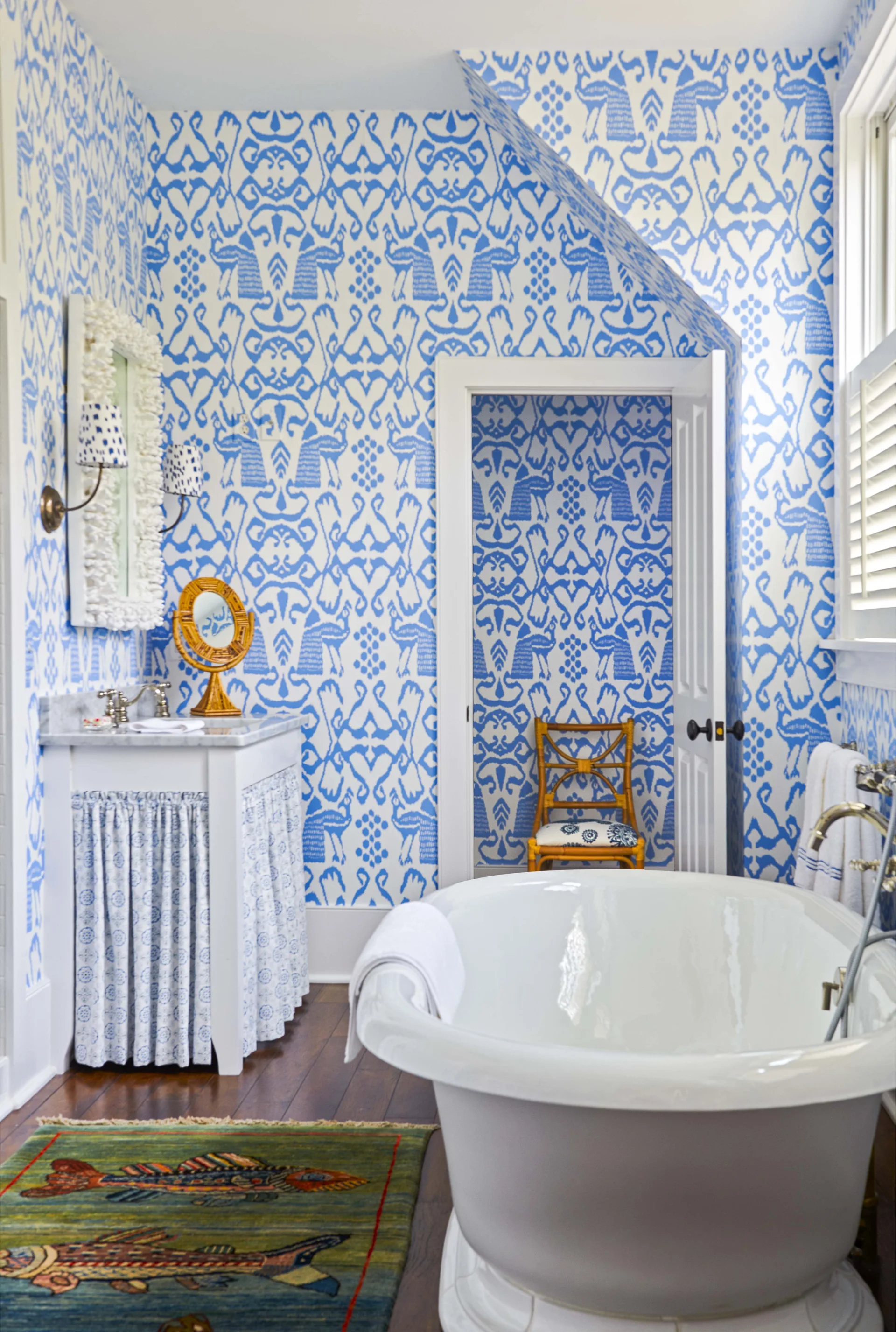 A bathroom features bold blue and white patterned wallpaper, a white freestanding bathtub, and a washstand vanity with a fabric skirt.