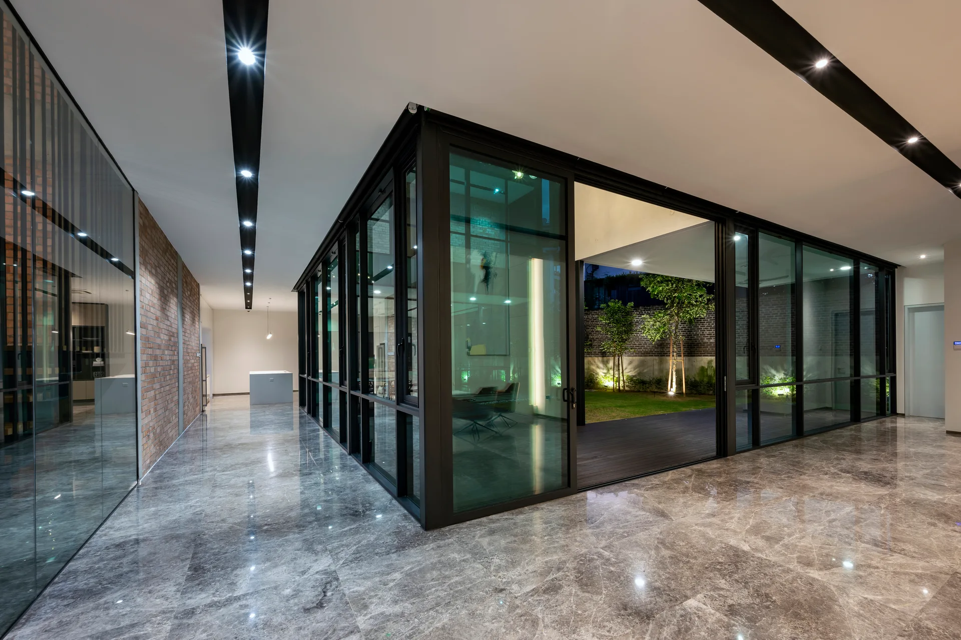 A modern interior corridor with polished gray marble floors, an exposed brick wall, and black-framed glass walls facing an outdoor courtyard.