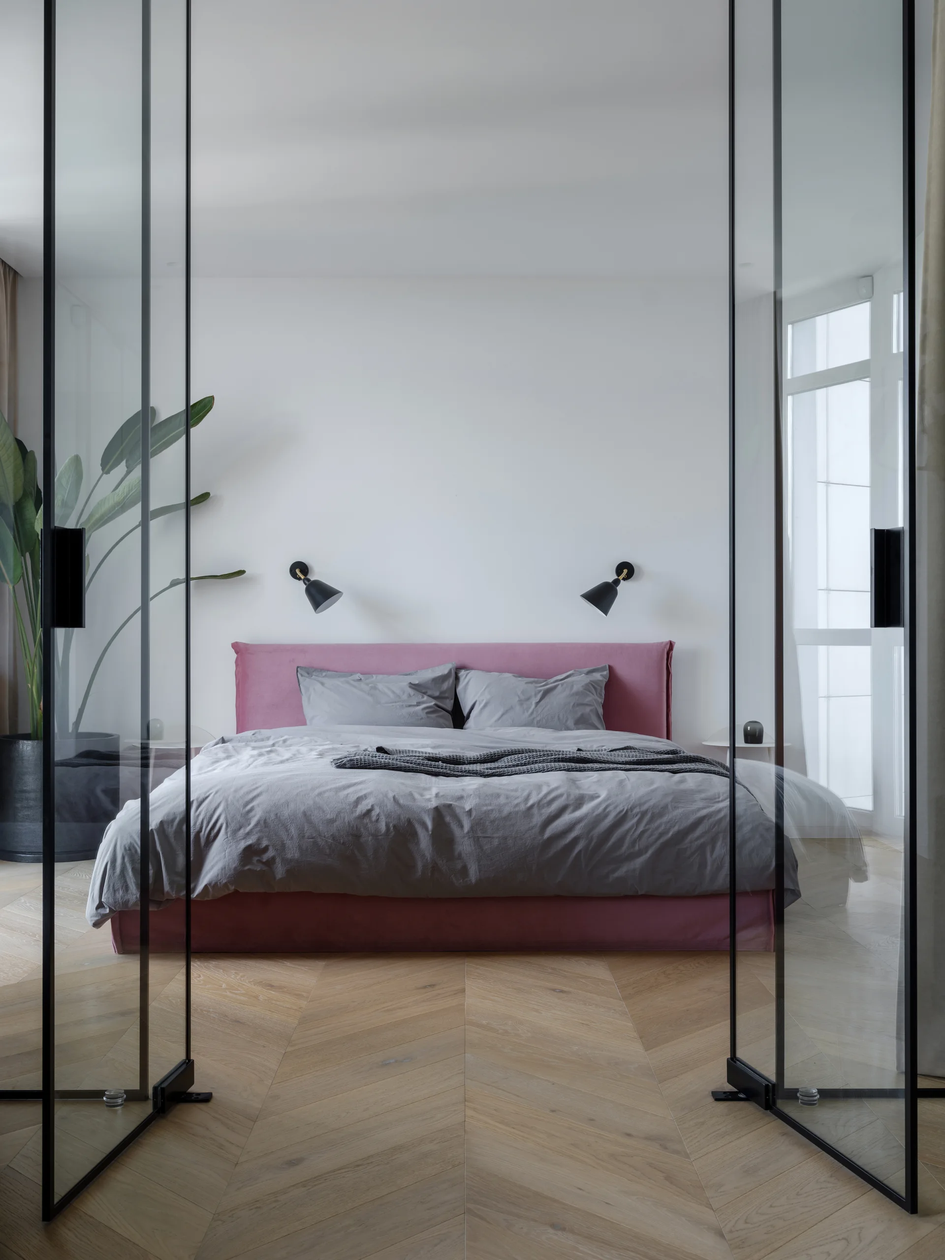 A modern bedroom with a pink upholstered bed and grey bedding is framed by black metal glass partition doors on herringbone wood floors.