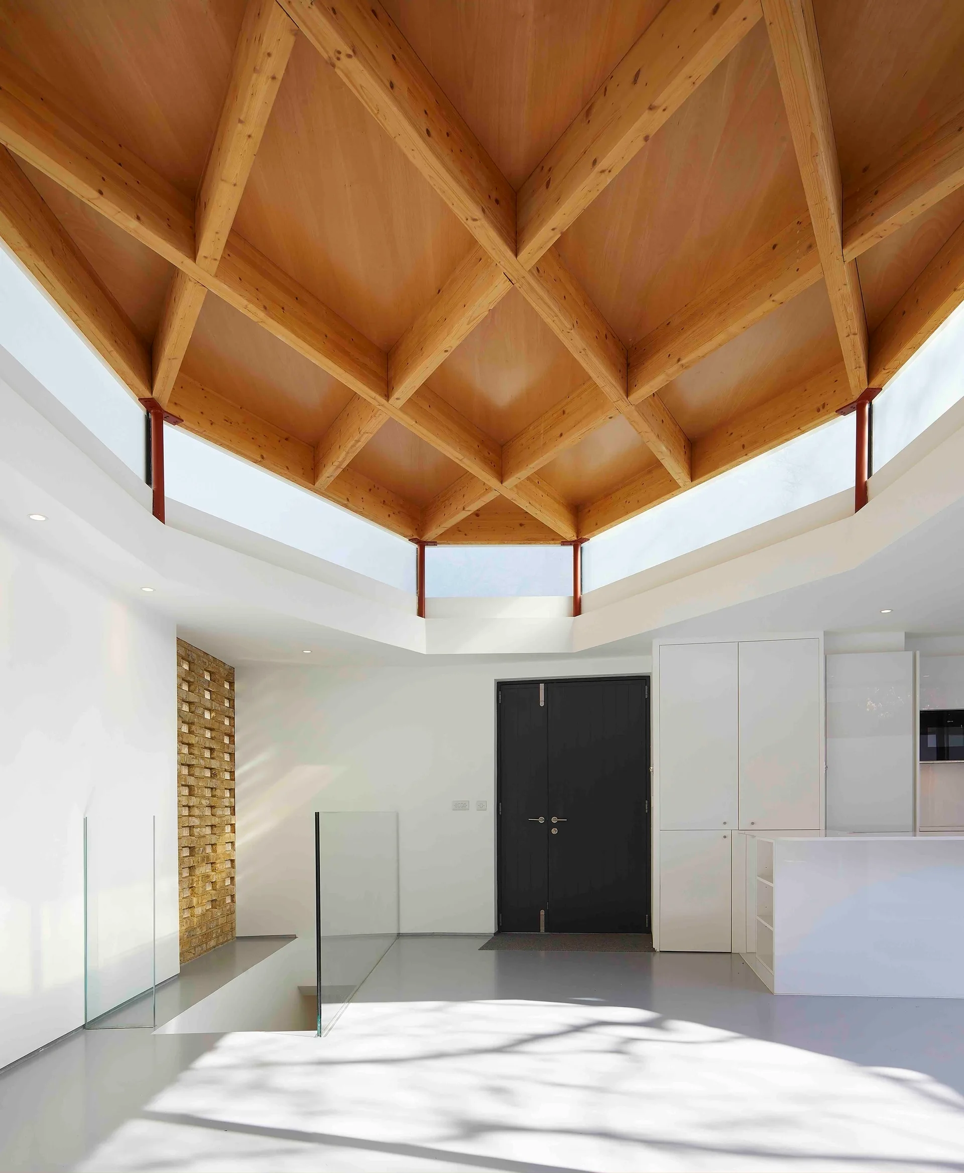 A modern entryway features a geometric wooden grid ceiling, white walls, clerestory windows, a black double door, and a glass staircase railing.