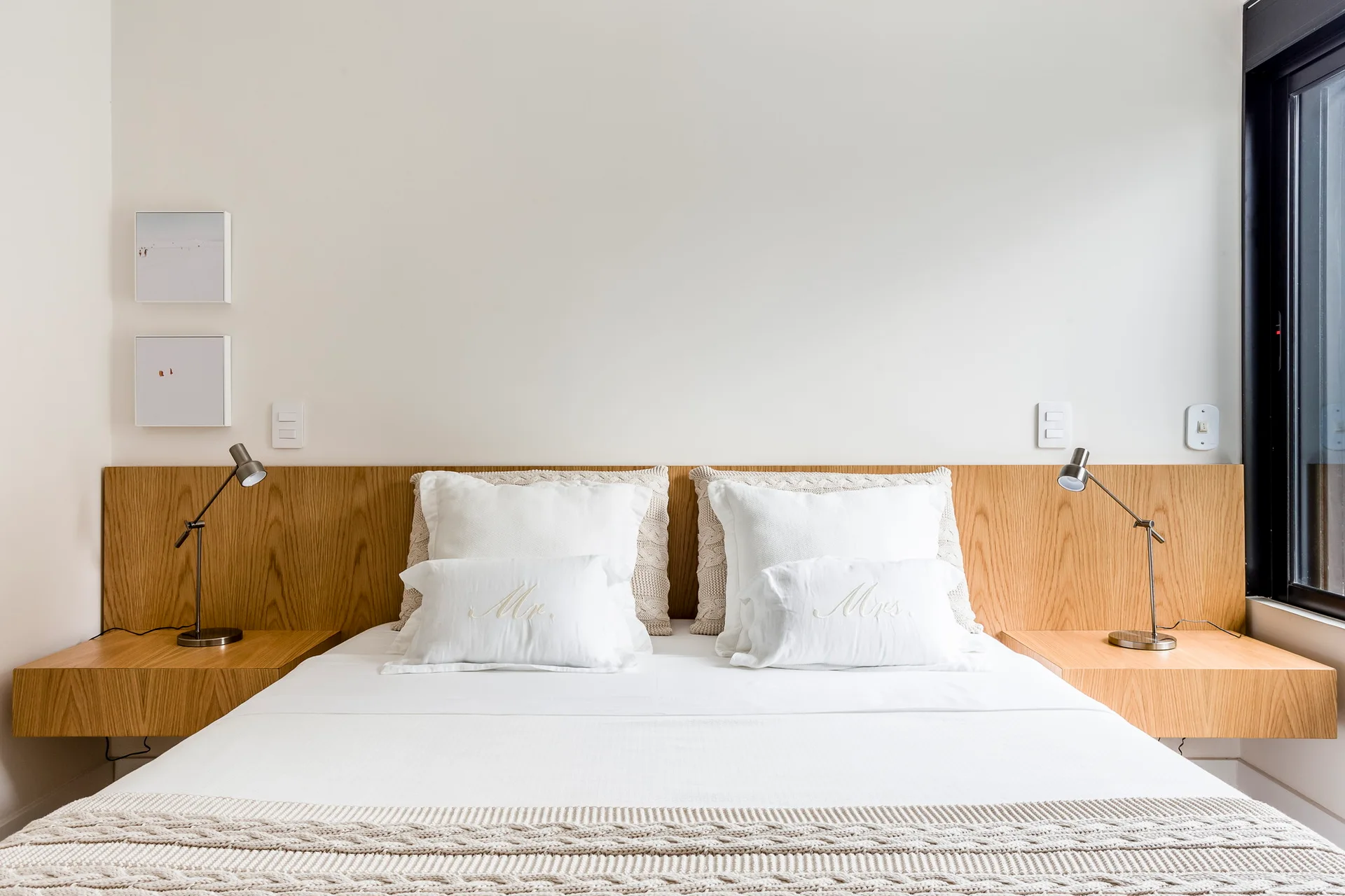A minimalist bedroom featuring a white bed set against a wide light wood headboard with integrated floating nightstands and bedside lamps.