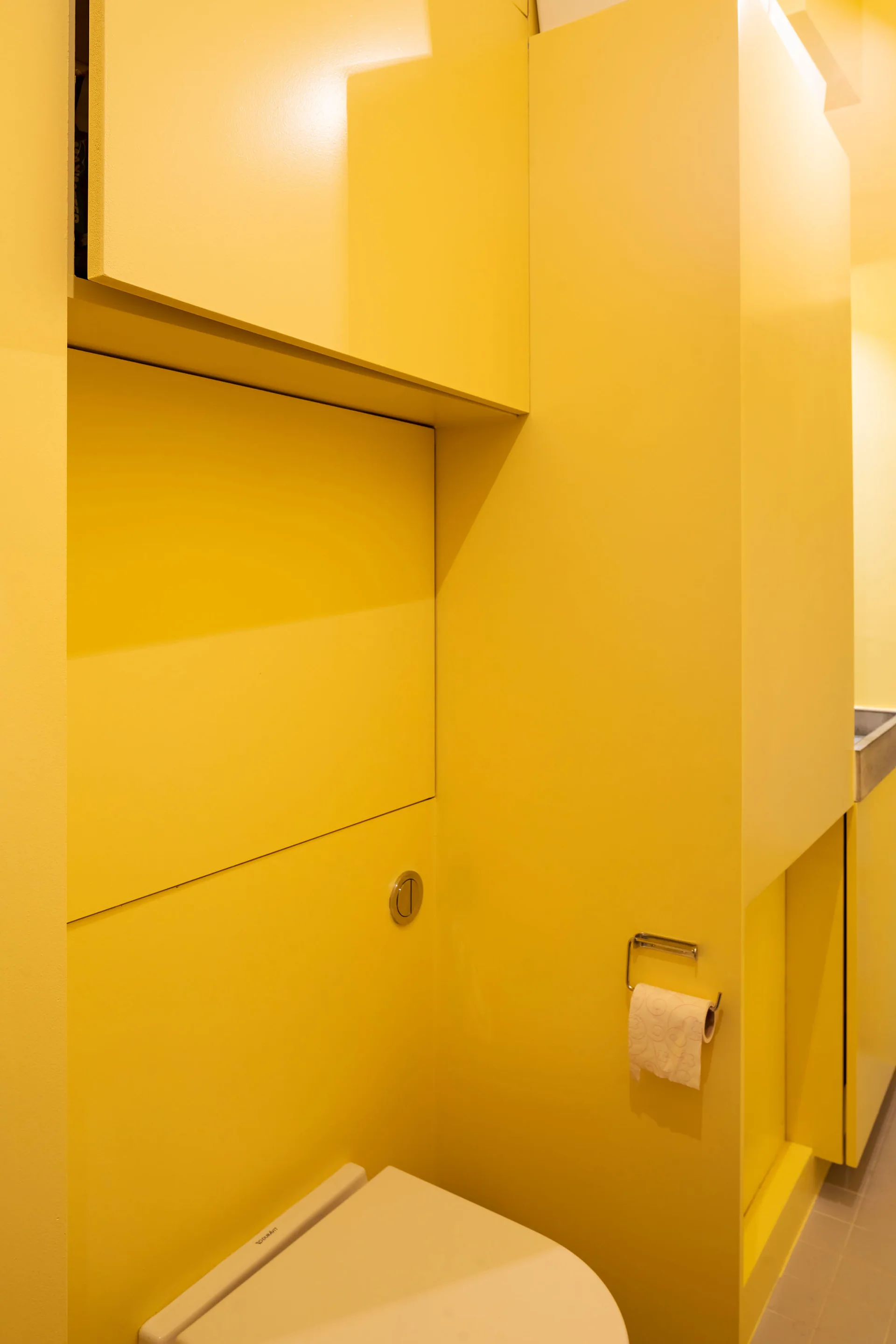 A modern yellow powder room featuring built-in satin cabinets, a white toilet seat, and a wall-mounted chrome flush button.