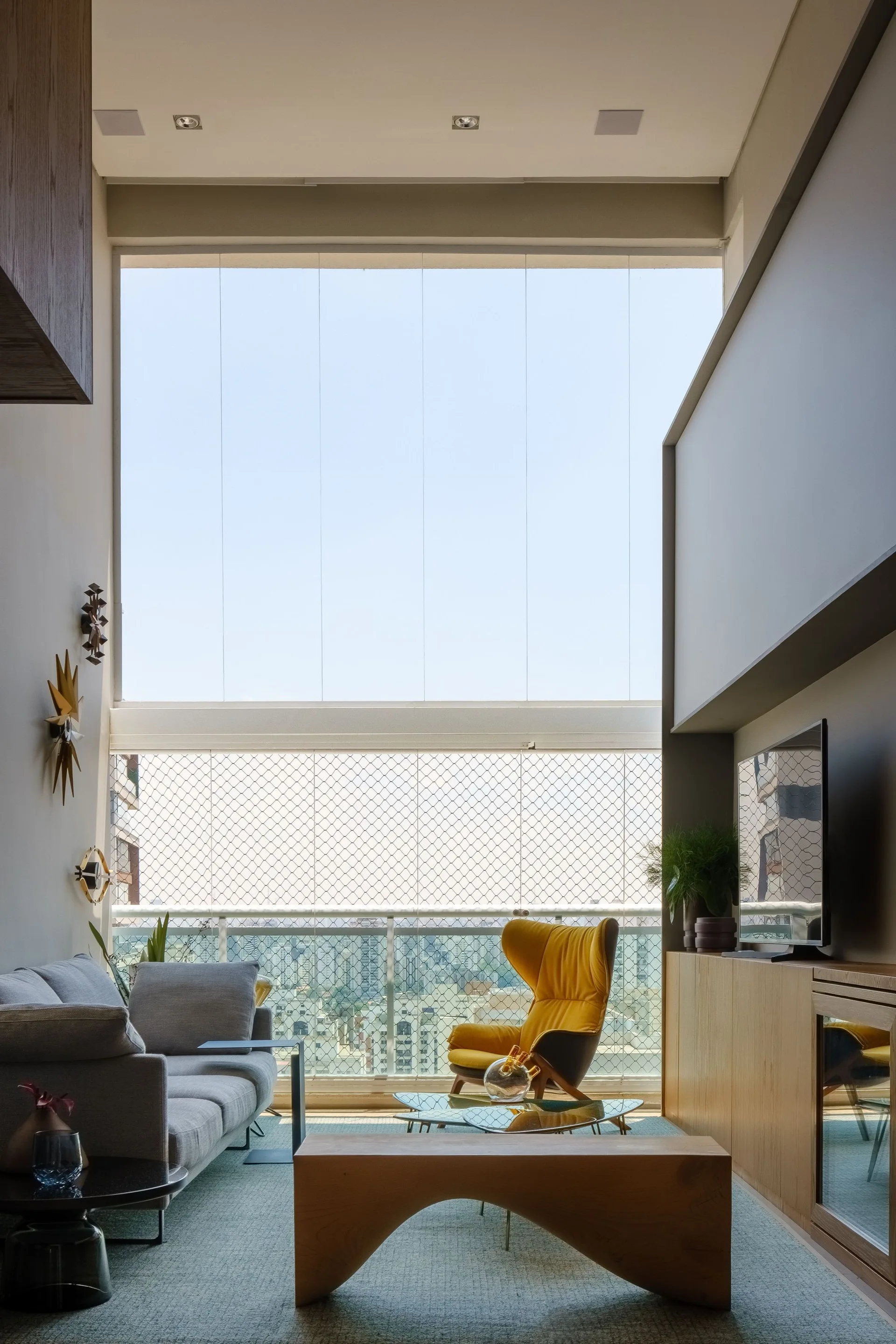 A light-filled living room features a grey sofa, a bright yellow winged armchair, a low wooden arched bench, and a large window opening to a balcony.