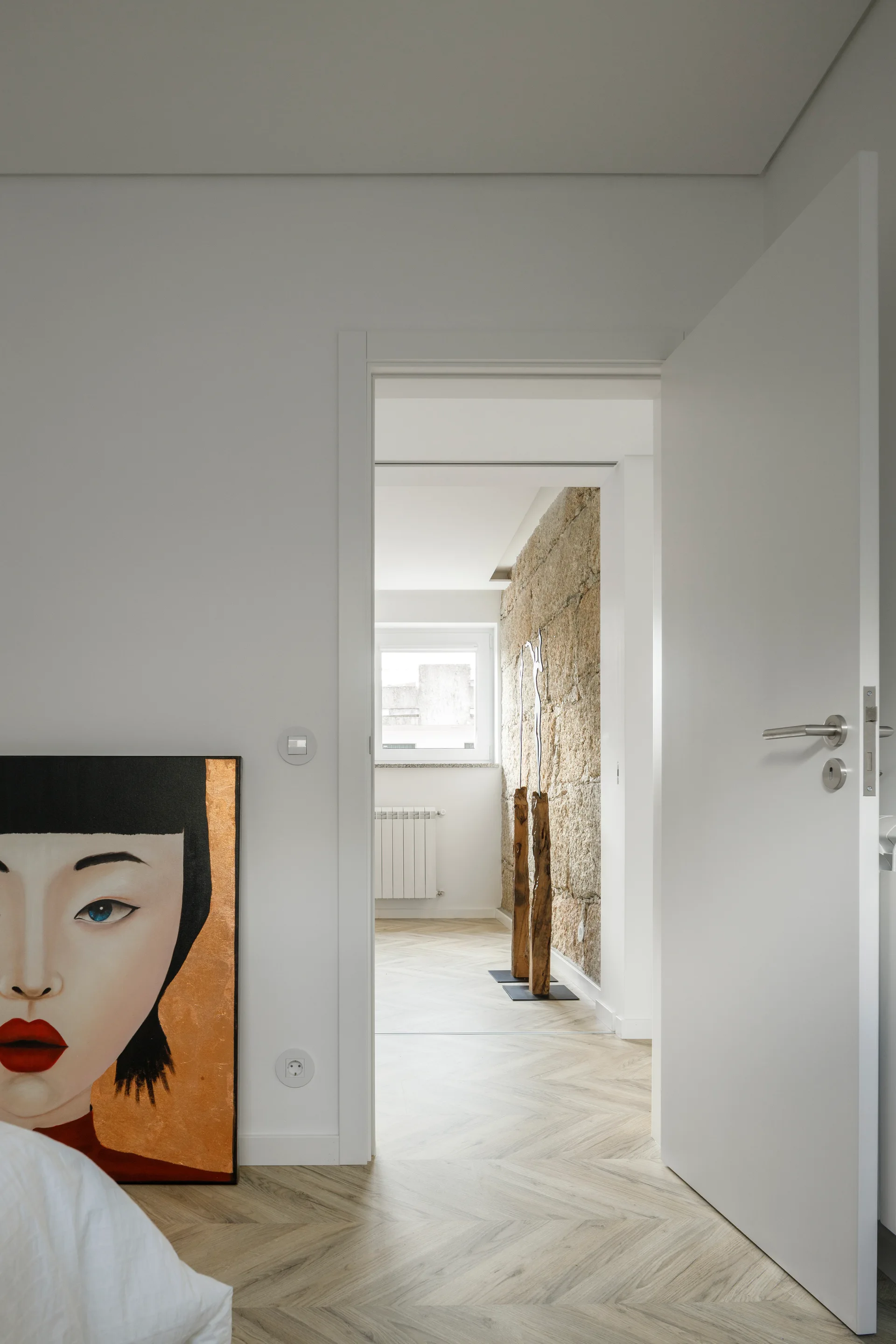 An open white door reveals a hallway featuring light wood herringbone floors, a textured stone accent wall, and a tall decorative wood sculpture.
