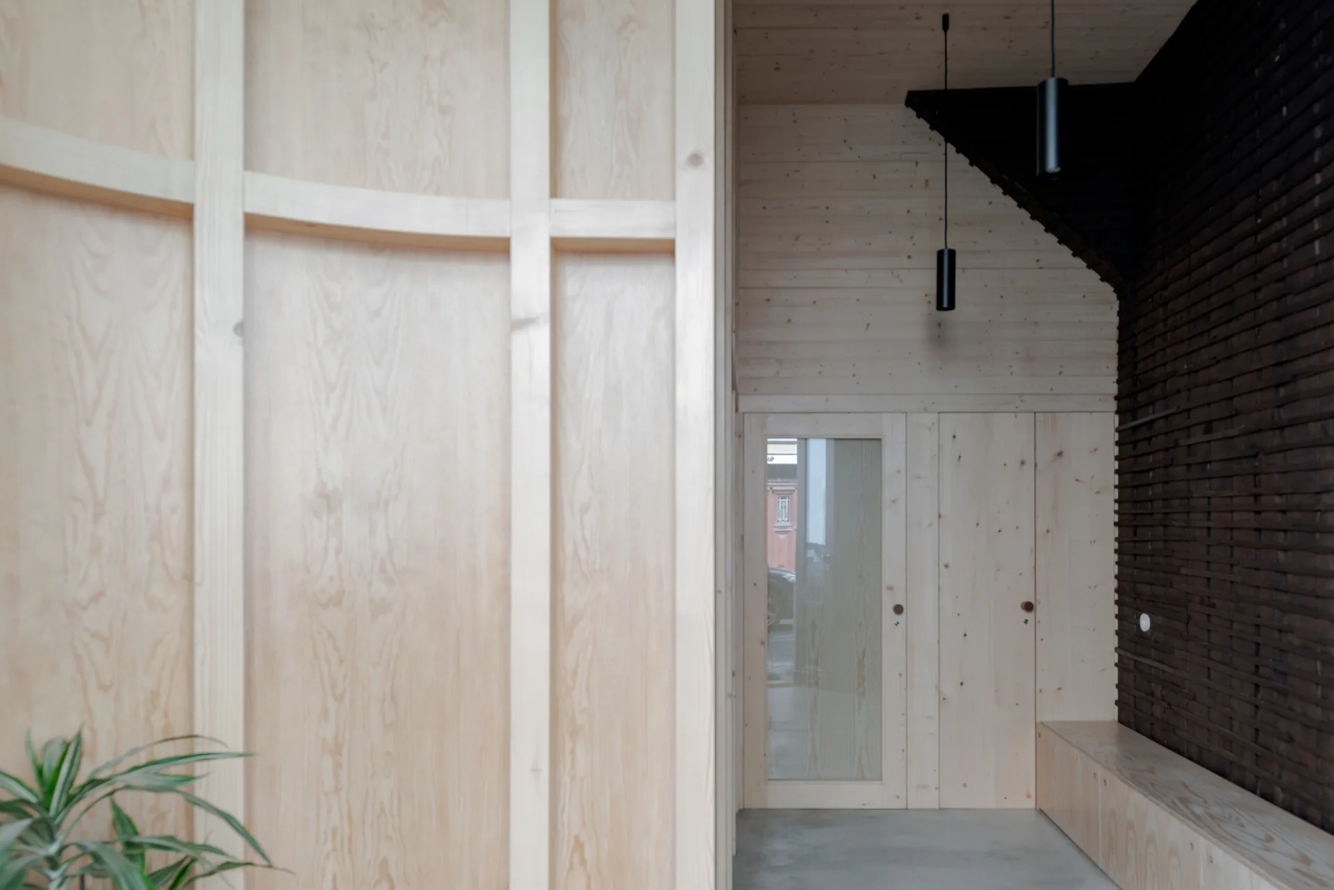 A minimalist entryway featuring light wood paneled walls, a built-in bench, dark brick wall cladding, and cylindrical black pendant lights.