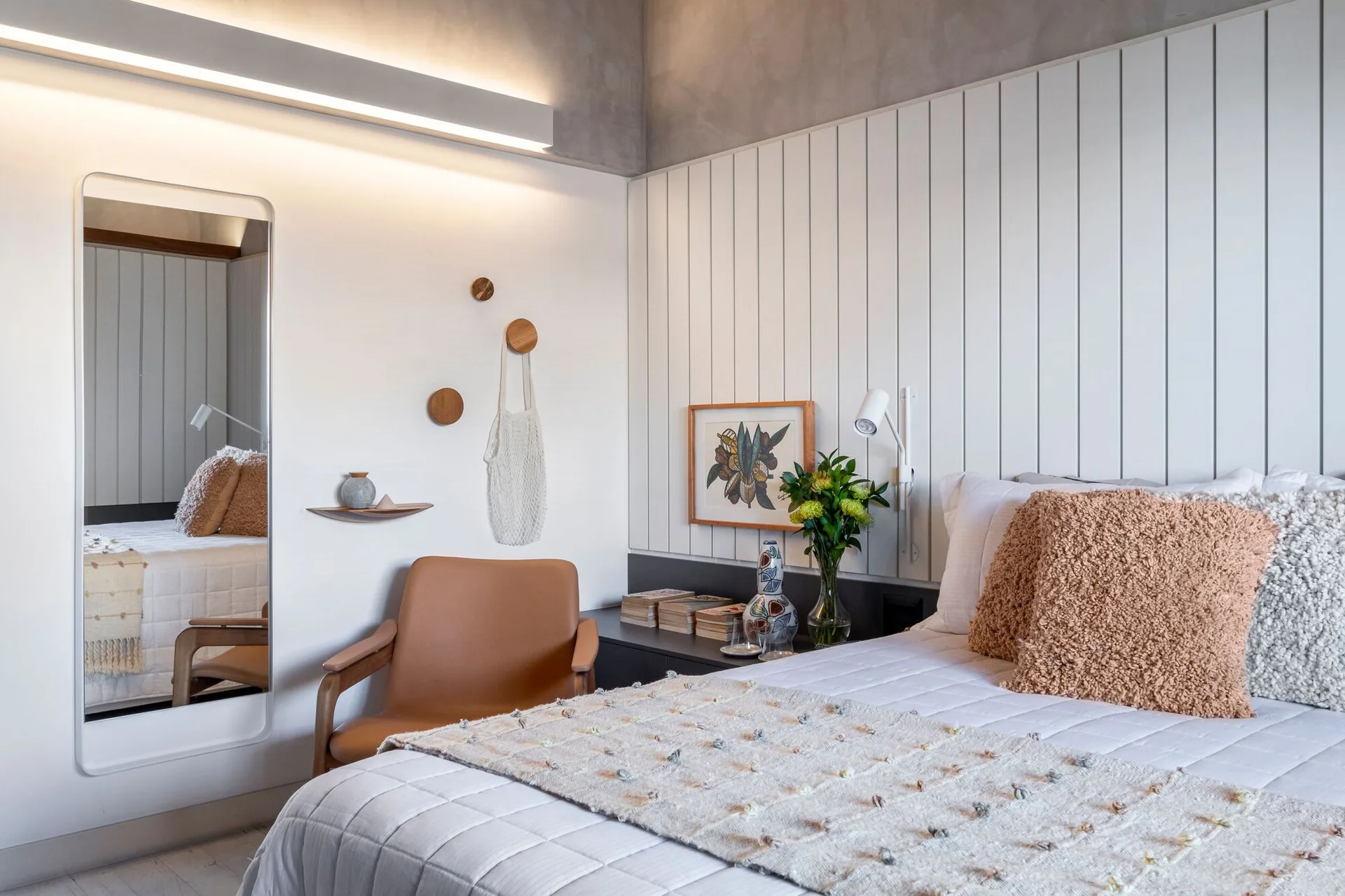 A modern bedroom features a bed with textured pillows, a white vertical wood slat wall, a brown leather armchair, and a tall rectangular wall mirror.