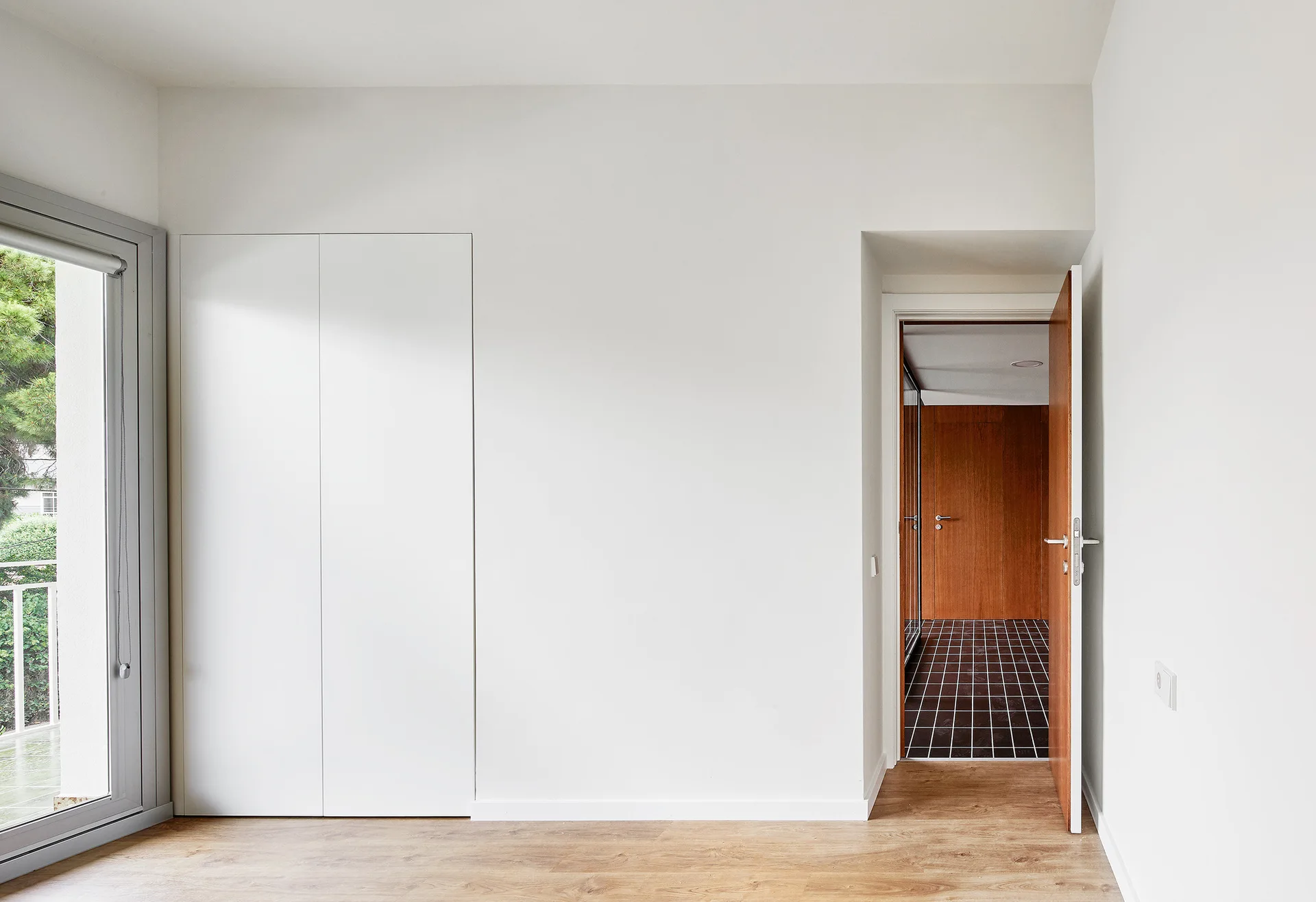 A minimalist white bedroom featuring flush-mount closet doors, wood flooring, and an open doorway leading to a wood-paneled room with black tiled floors.