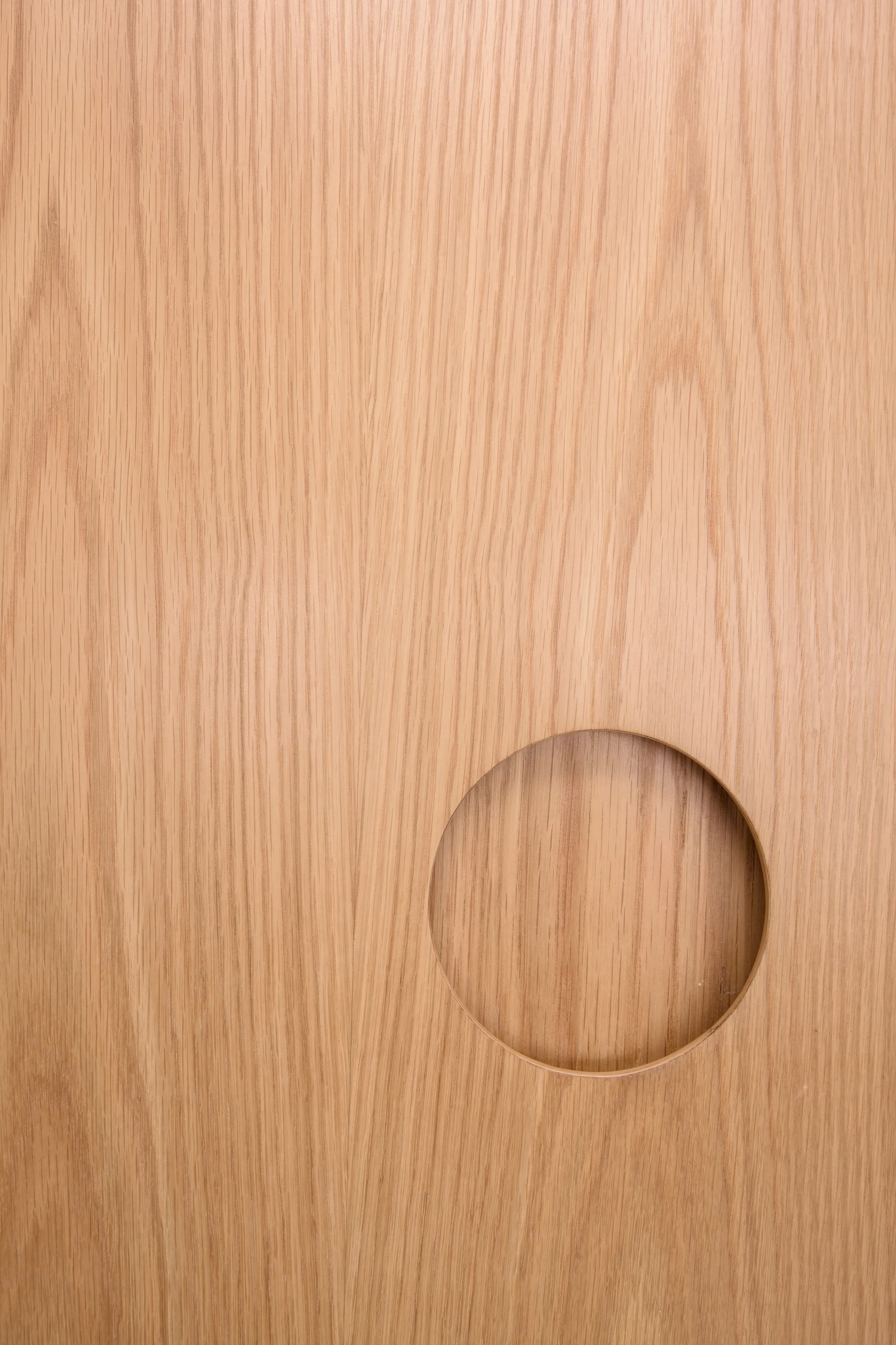 A close-up of a light oak wood panel with a vertical grain and a circular recessed cutout handle near the bottom right.