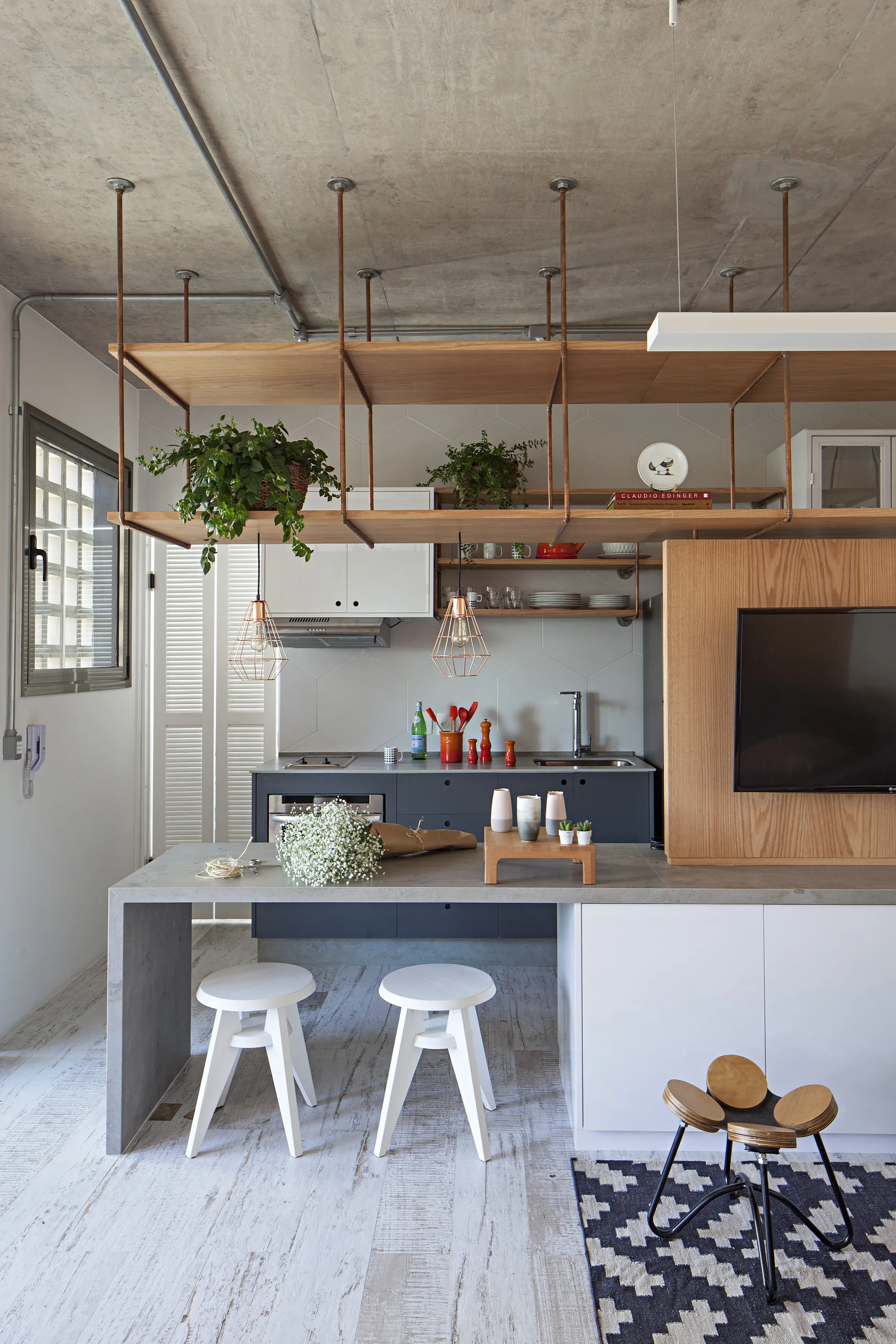 A contemporary industrial kitchen featuring a concrete island with white wood stools below suspended shelving made of wood planks and copper piping.