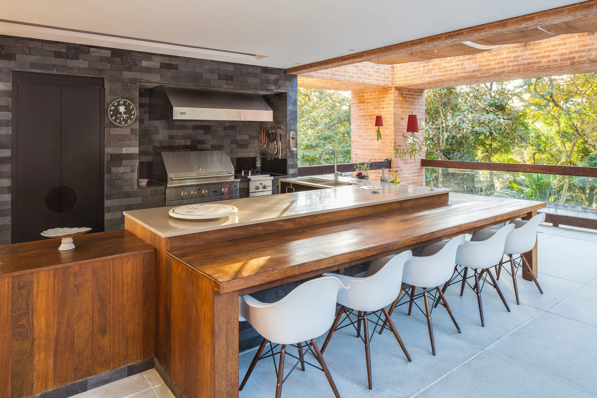 A modern covered terrace features a stone outdoor kitchen with a stainless steel grill, a wood island with white bar stools, and views of green trees.