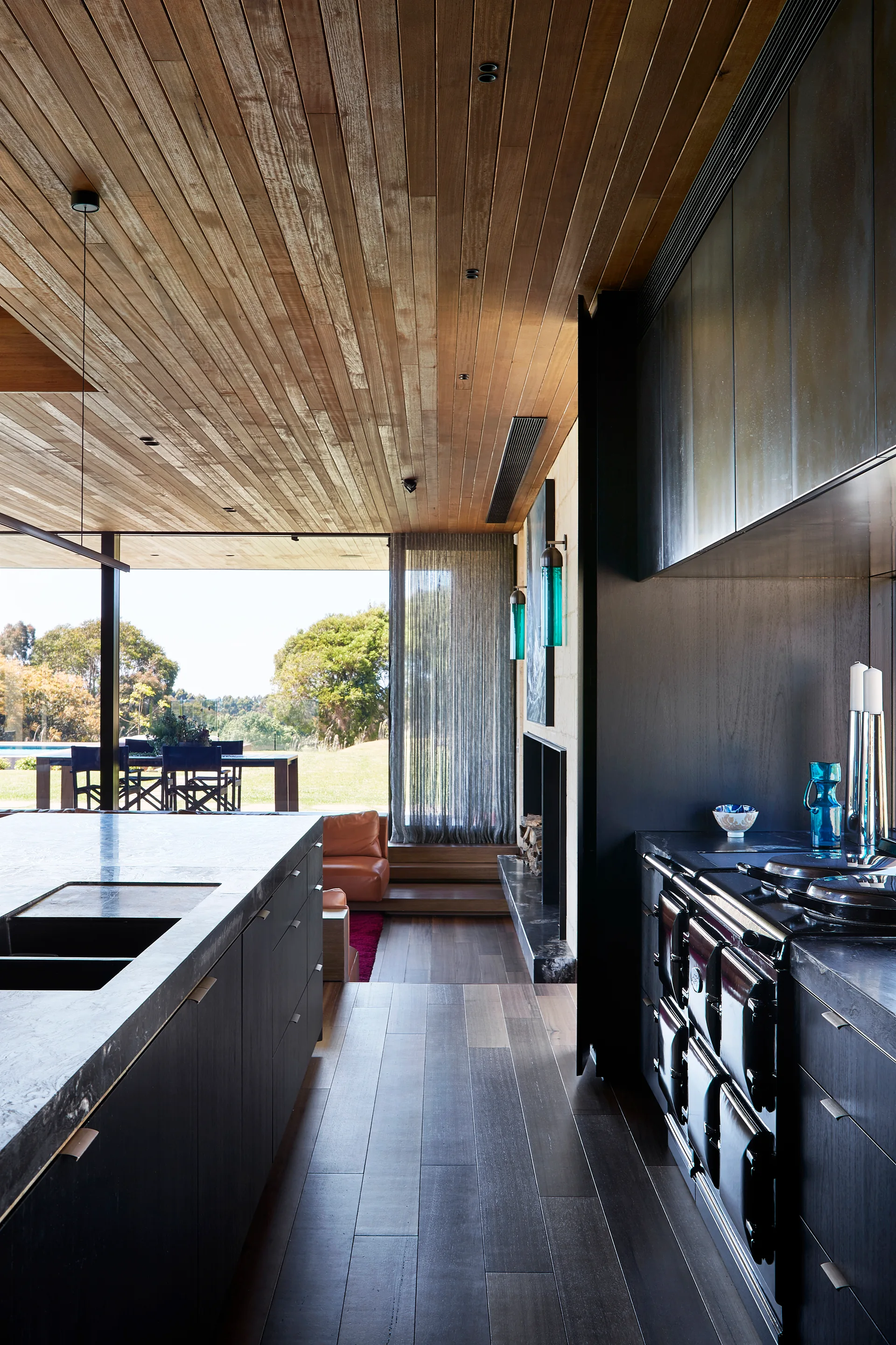 A modern kitchen featuring dark wood cabinetry, a large stone-topped island, and a warm wood-planked ceiling overlooking an outdoor patio.