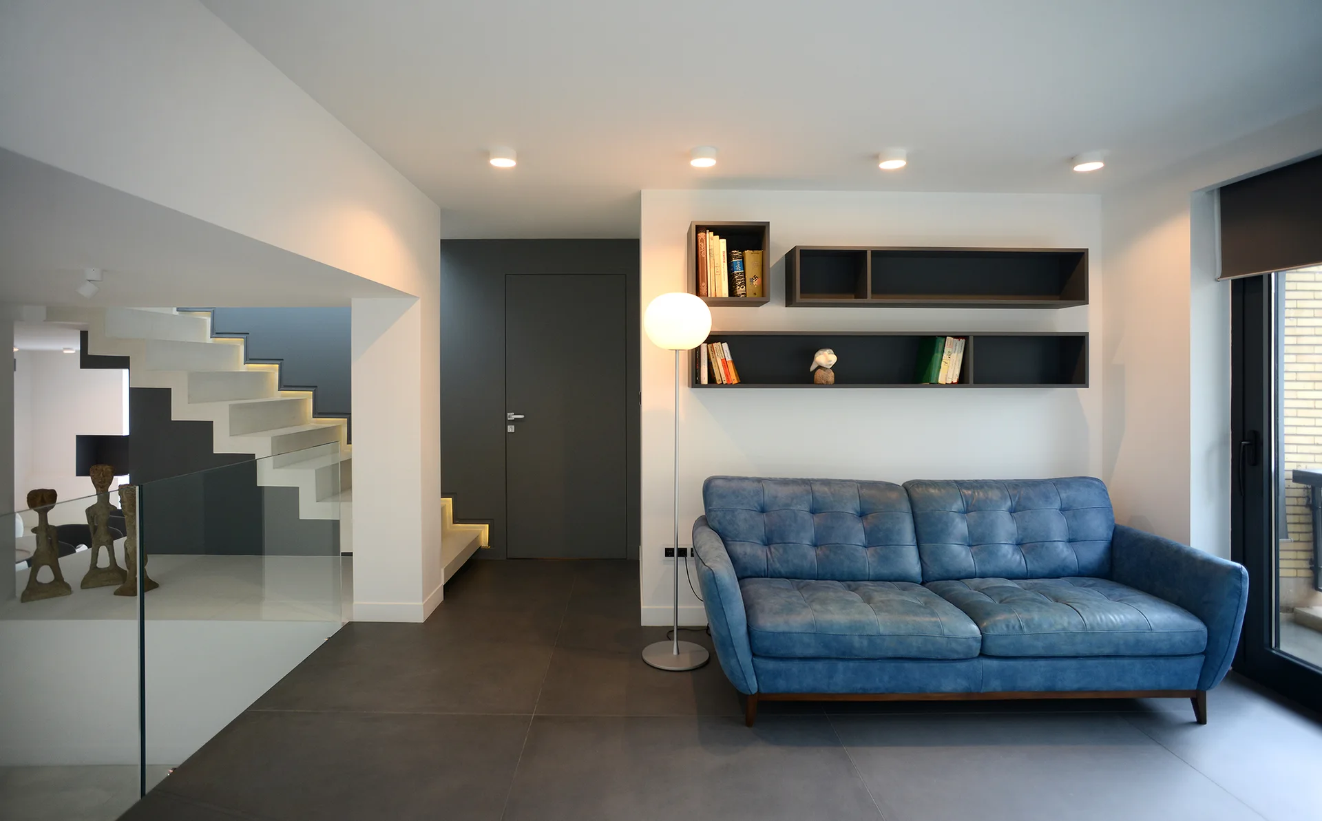 A contemporary living room featuring a blue tufted leather sofa, dark wall-mounted shelf boxes, and a lighted floating staircase.