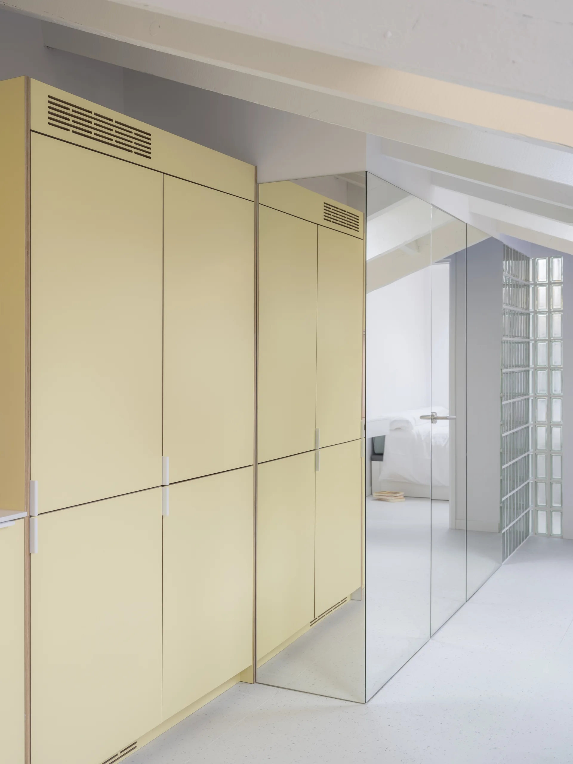 Matte pale yellow cabinets stand beside a full-length mirror and a translucent glass block wall under white sloped ceiling beams.