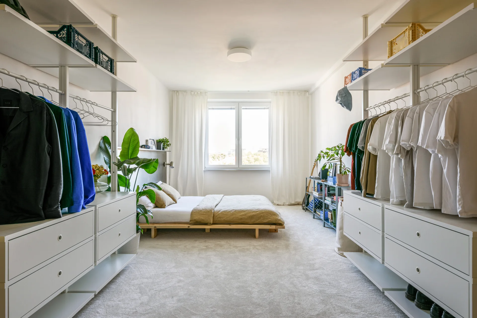 A minimalist bedroom features a central low wooden platform bed flanked by symmetrical white open wardrobe systems under a bright window.