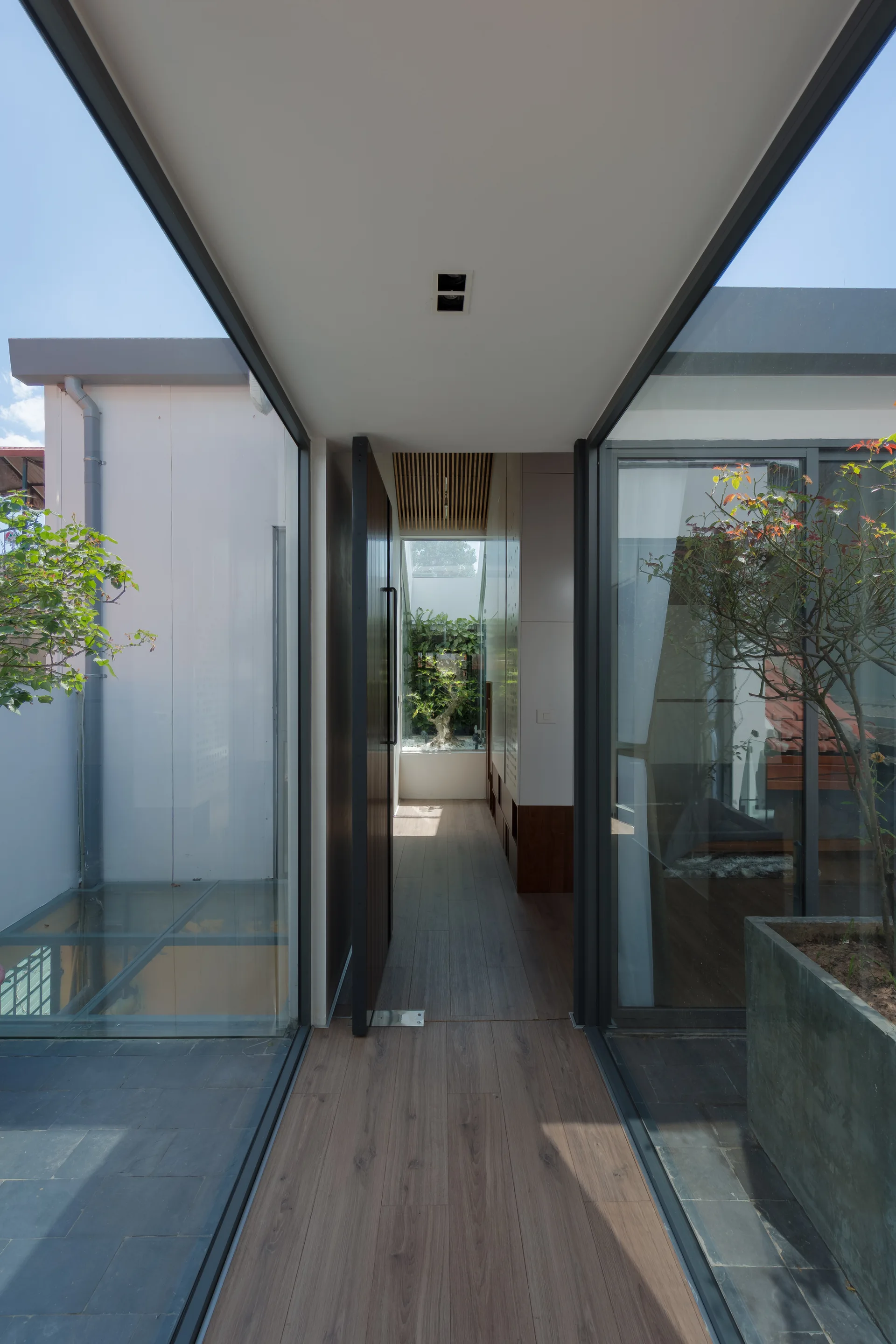 A modern minimalist hallway features floor-to-ceiling glass walls, wood plank flooring, and a view into a small interior courtyard under a skylight.