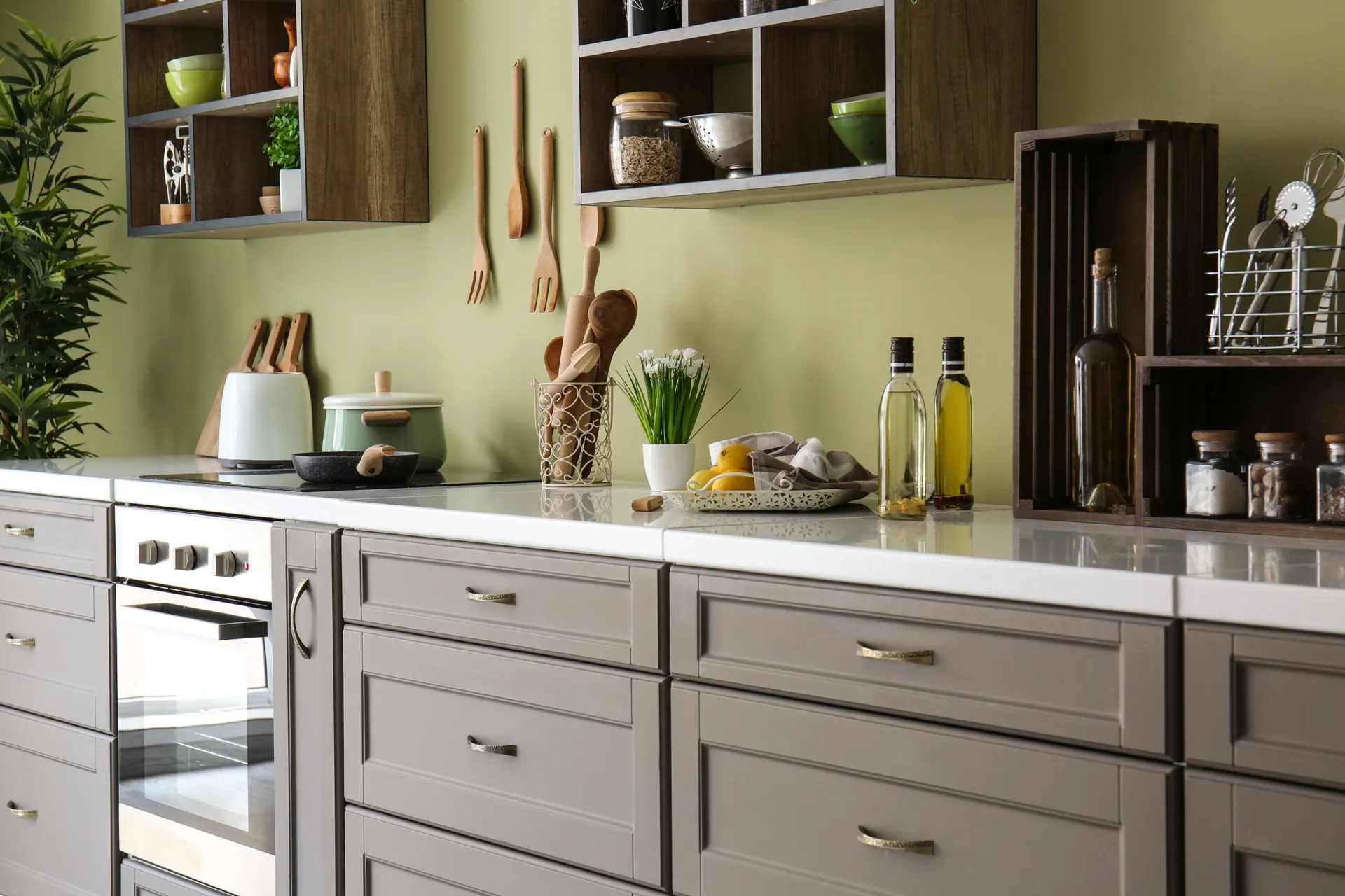 A kitchen features grey Shaker-style cabinets, a white countertop, an olive green wall, and dark wood open shelving.
