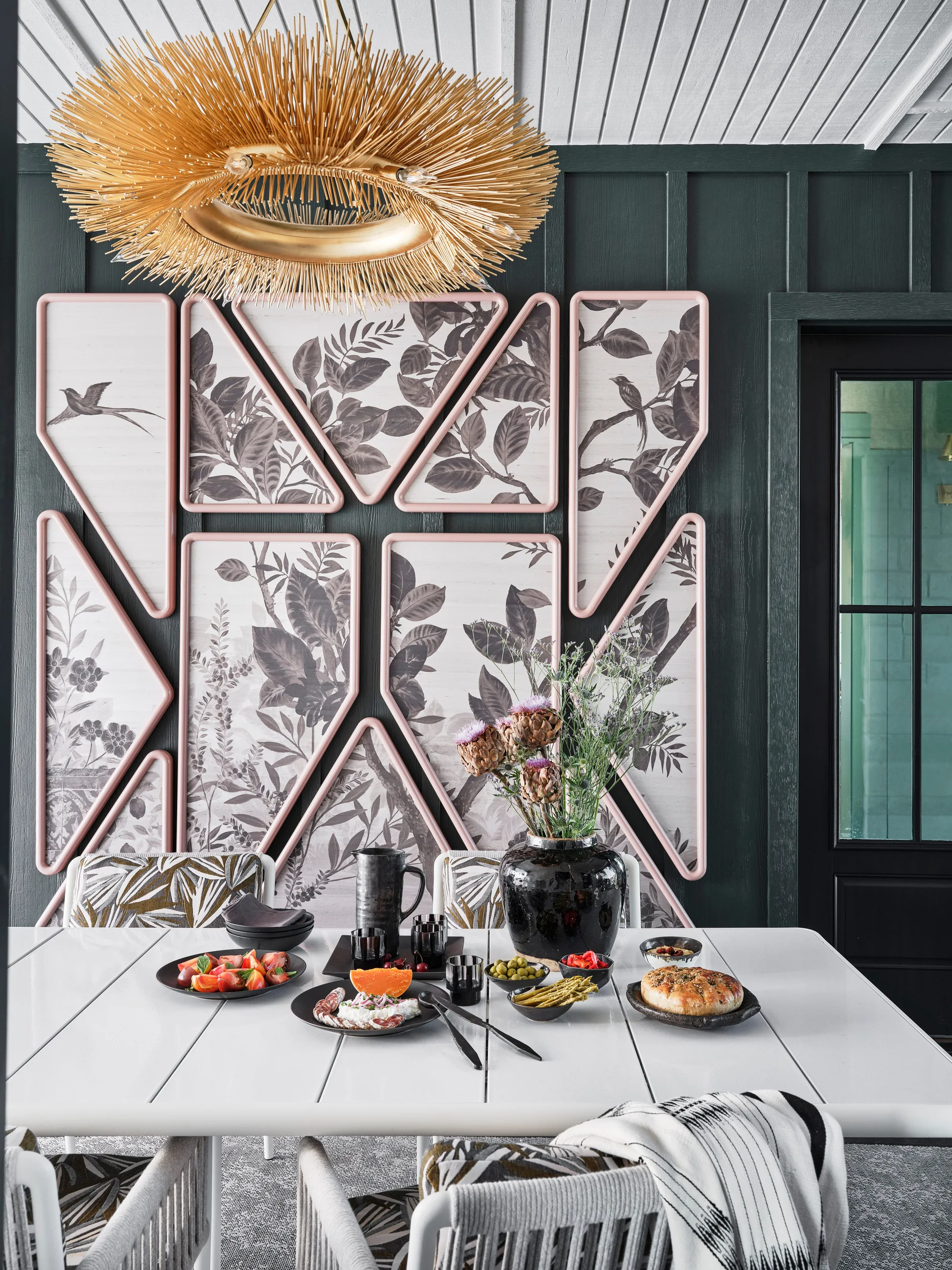A white dining table set with food sits in front of a dark green paneled wall decorated with geometric pink-framed botanical art.