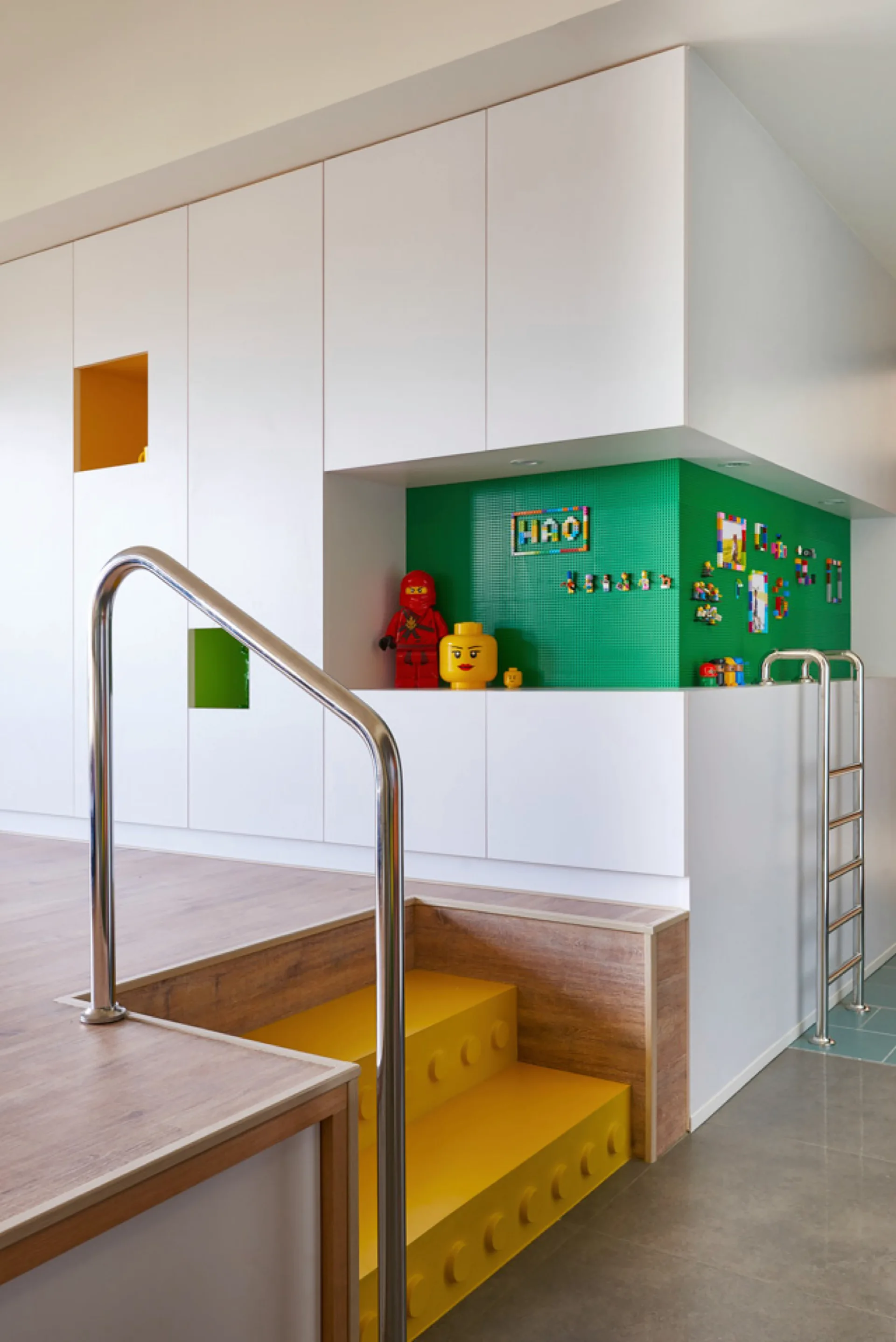 White built-in cabinetry sits above a wood-toned platform with integrated yellow steps and a green Lego-textured accent wall.