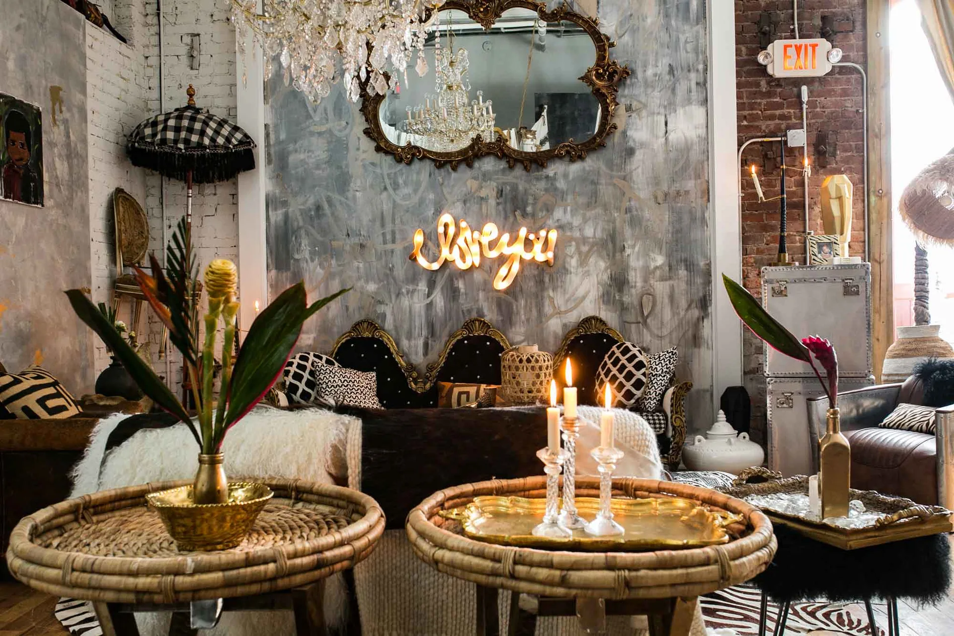 An eclectic living room features a black velvet sofa beneath a neon sign, paired with two round woven coffee tables and a crystal chandelier.