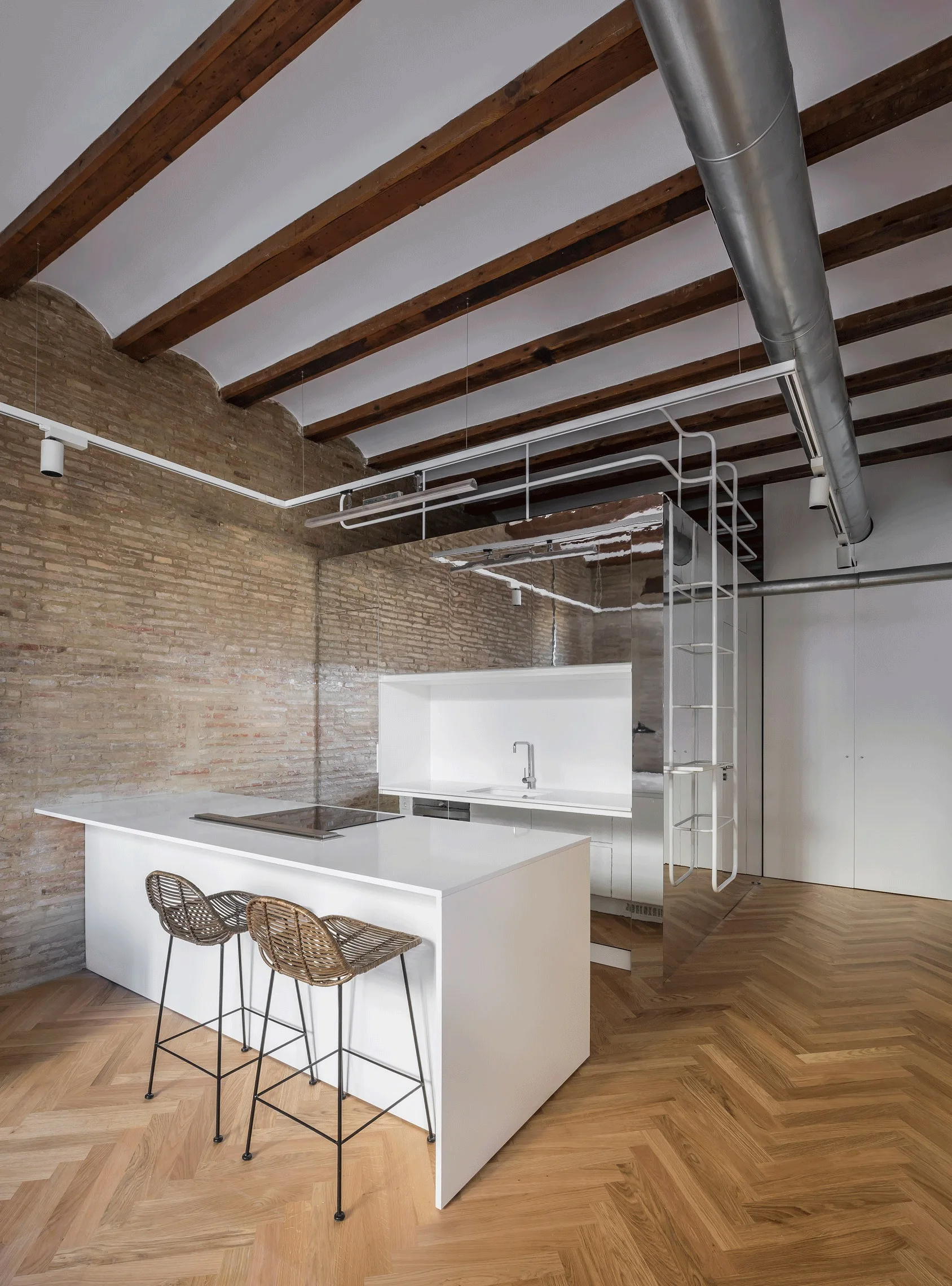 A minimalist kitchen featuring a white island with woven bar stools, an exposed brick wall, and a mirrored wall panel under exposed wooden ceiling beams.