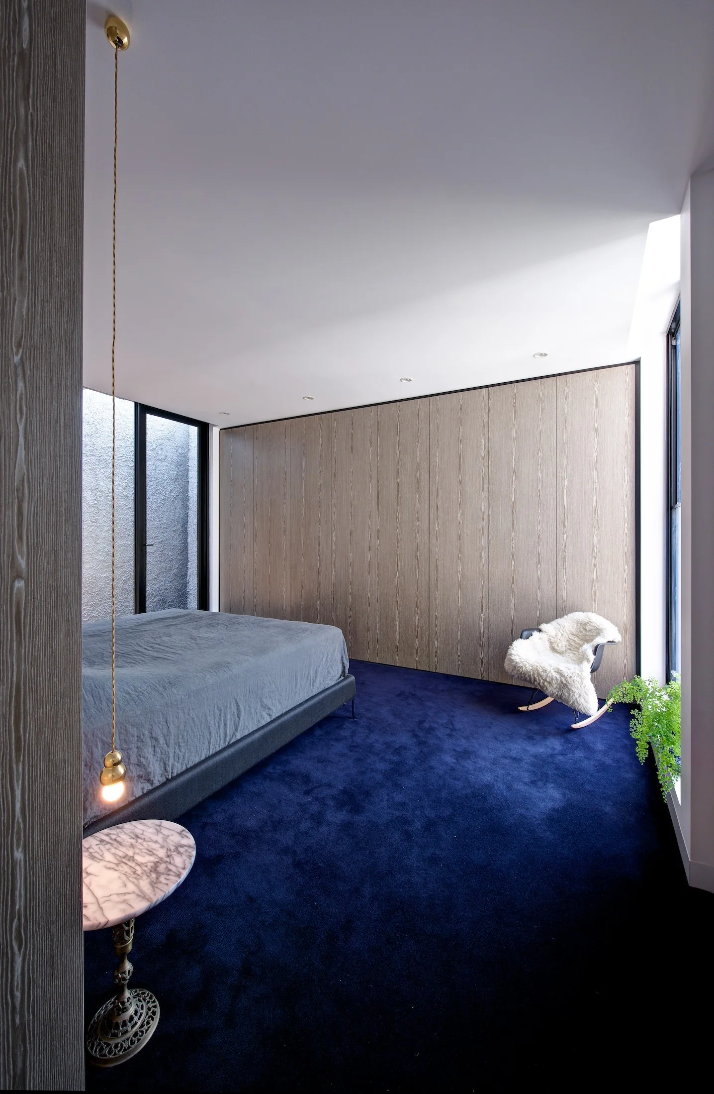 A contemporary bedroom features a bed with grey sheets on deep blue carpet next to a floor-to-ceiling wood paneled wall and a white rocking chair.