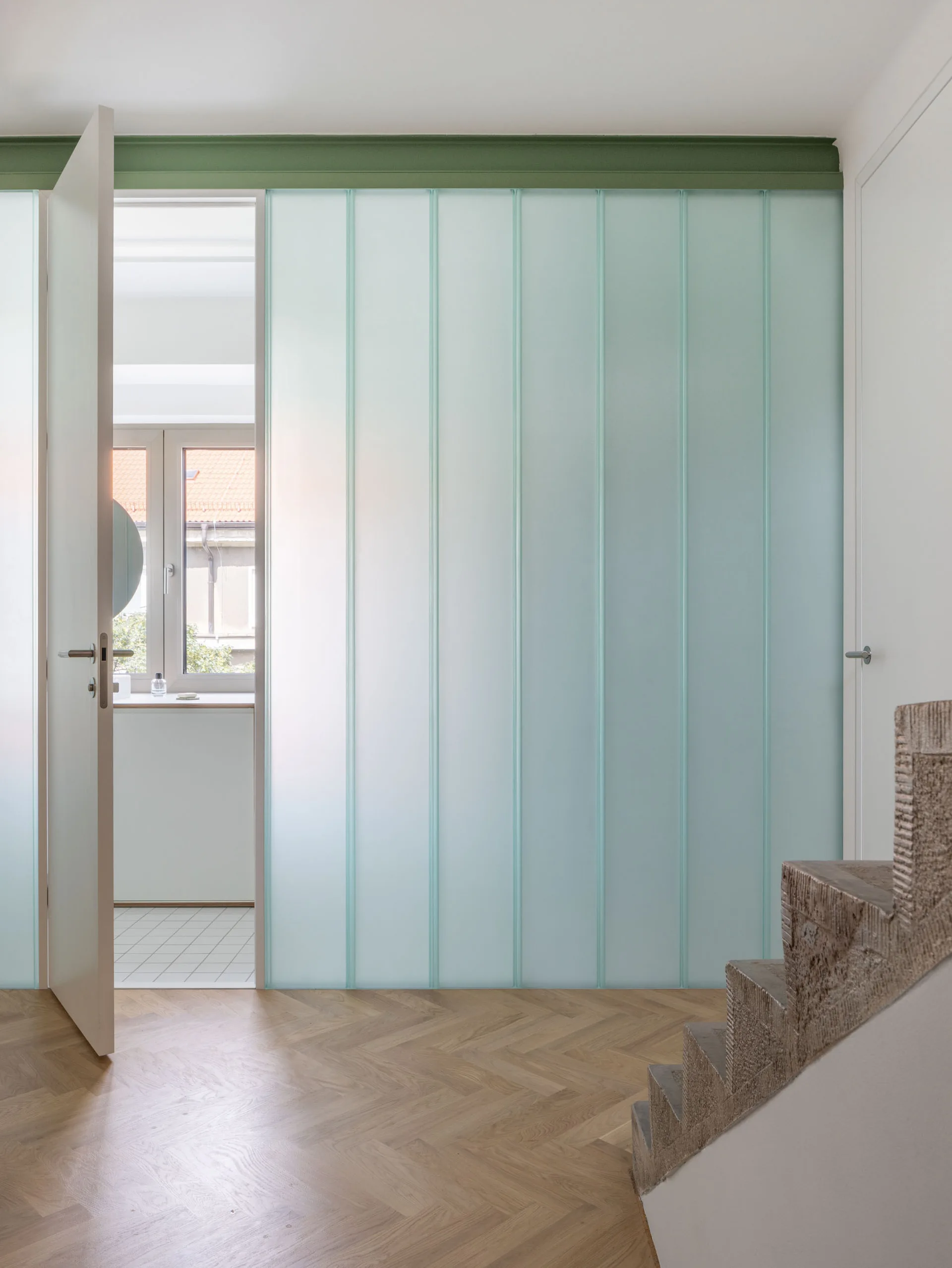 A contemporary hallway featuring a frosted glass partition wall, light wood herringbone flooring, and the edge of a textured stone staircase.