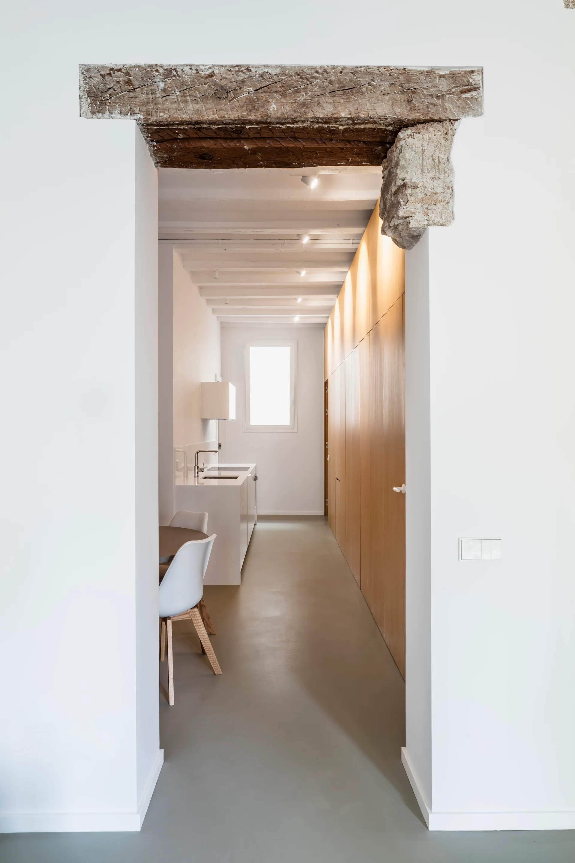 A view through a rustic wooden entryway reveals a minimalist kitchen with seamless wood cabinetry, a white island, and grey flooring.