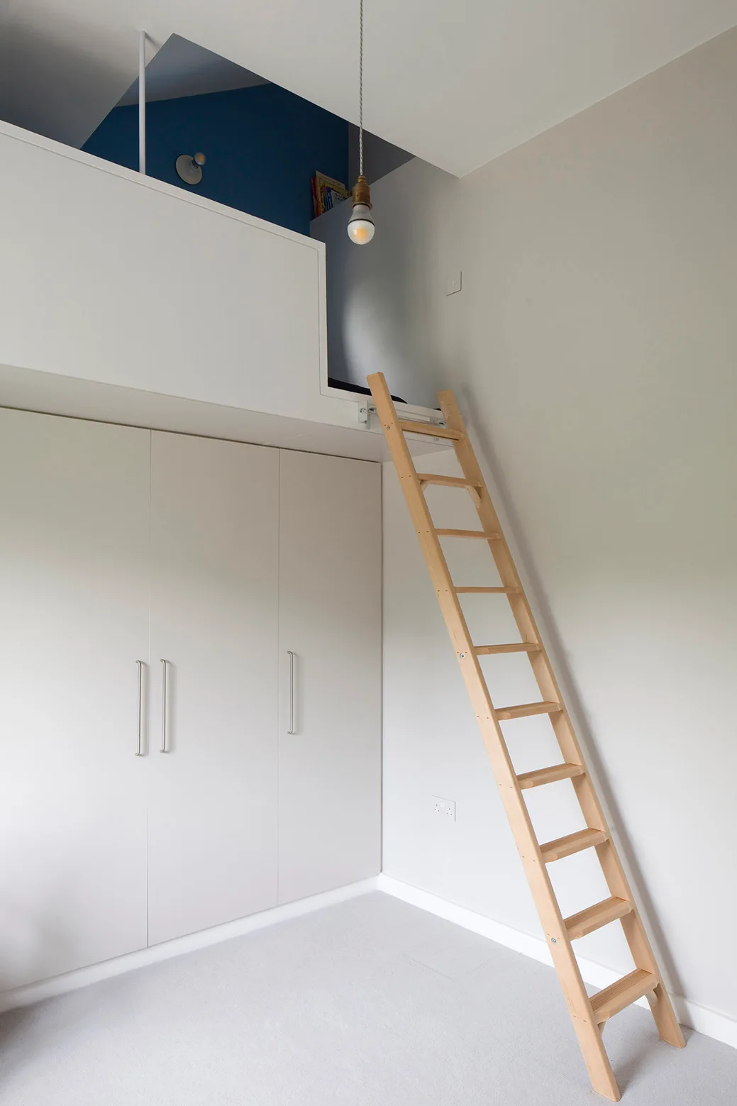 A light wood ladder leans against a built-in white wardrobe leading to a loft sleeping platform with a blue accent wall and hanging pendant light.