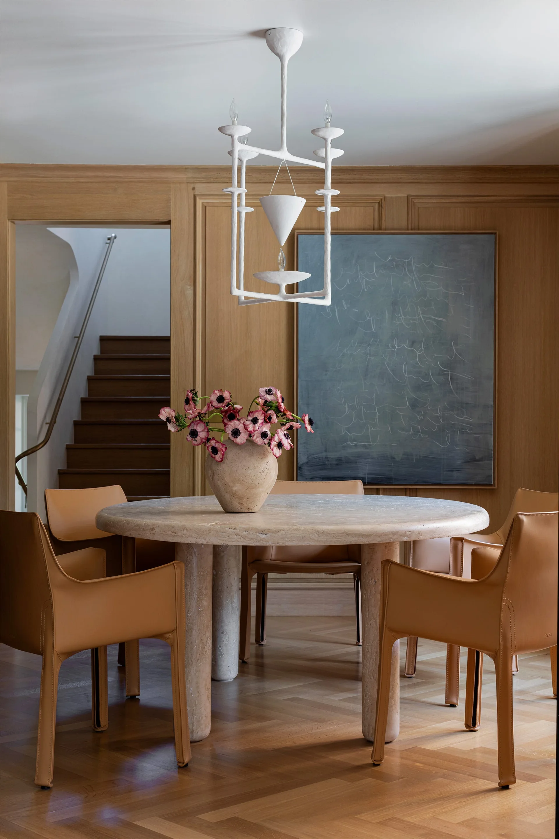 A round stone dining table surrounded by tan leather chairs is set against warm wood-paneled walls beneath a white sculptural chandelier.