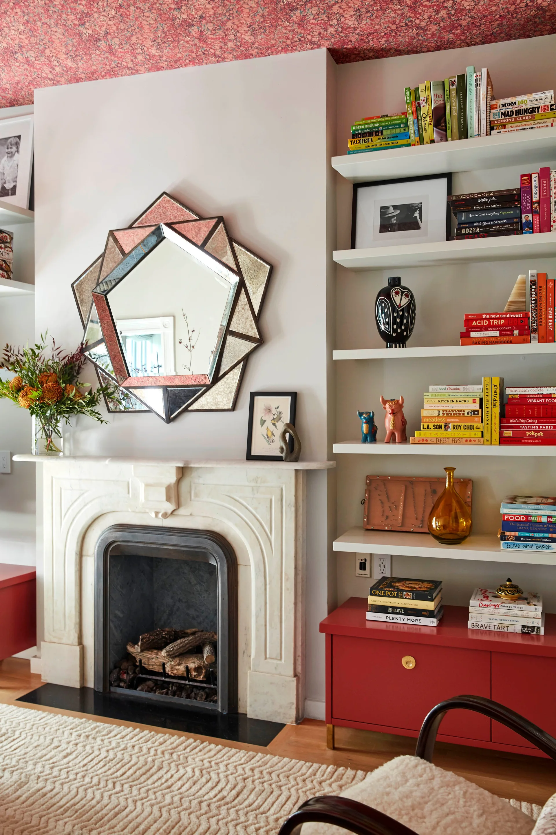 A marble fireplace with a geometric starburst mirror is flanked by white bookshelves and a red base cabinet under a patterned red ceiling.