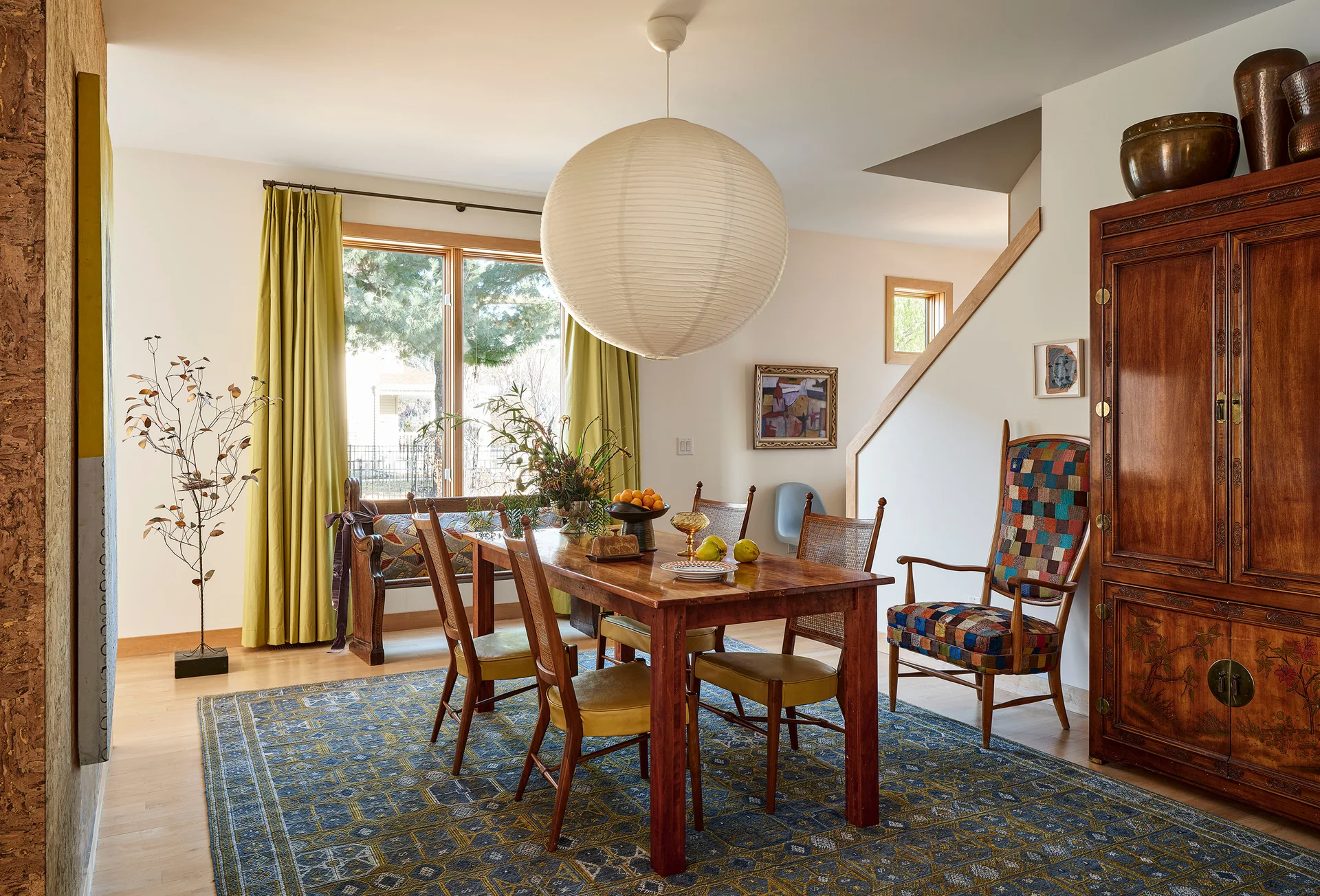 An eclectic dining room featuring a wooden table, cane-backed chairs, a patterned blue area rug, and an oversized paper globe pendant light.