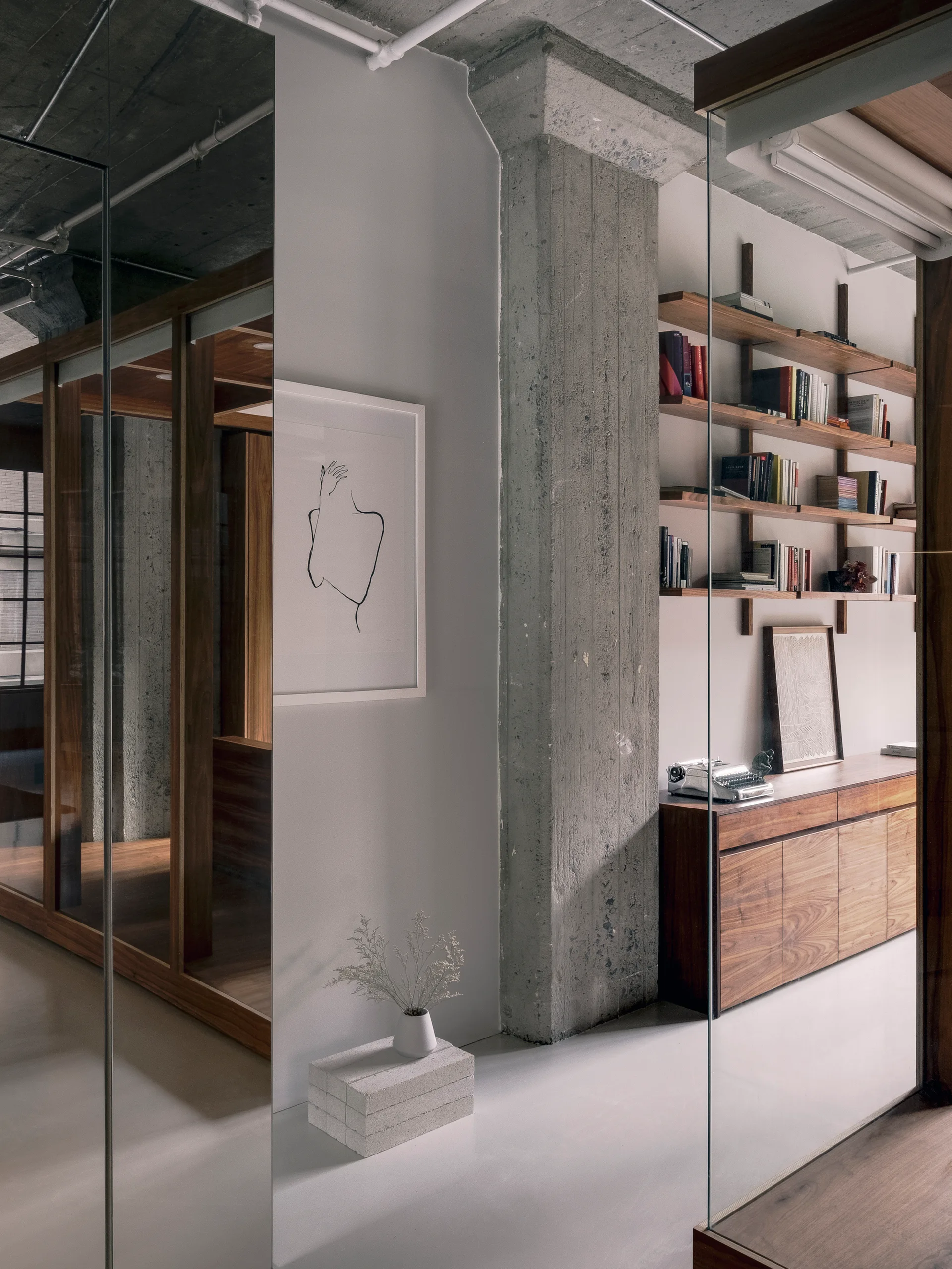 A modern library room featuring a prominent concrete pillar, glass partition walls, floating wooden shelves, and a matching wood cabinet.