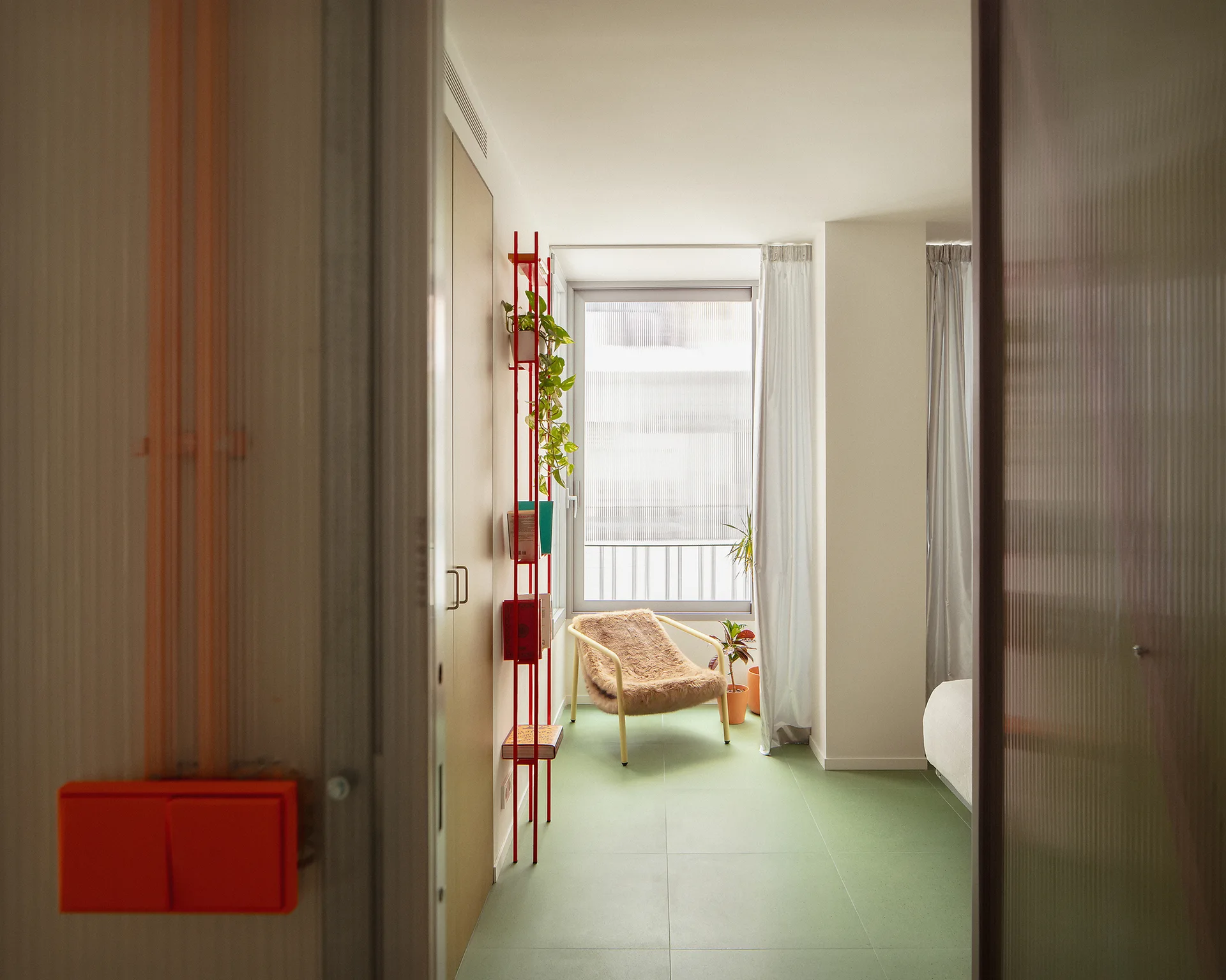 A minimalist bedroom viewed through a doorway, displaying light green tiled floors, a red vertical shelf, and a furry armchair near a window.