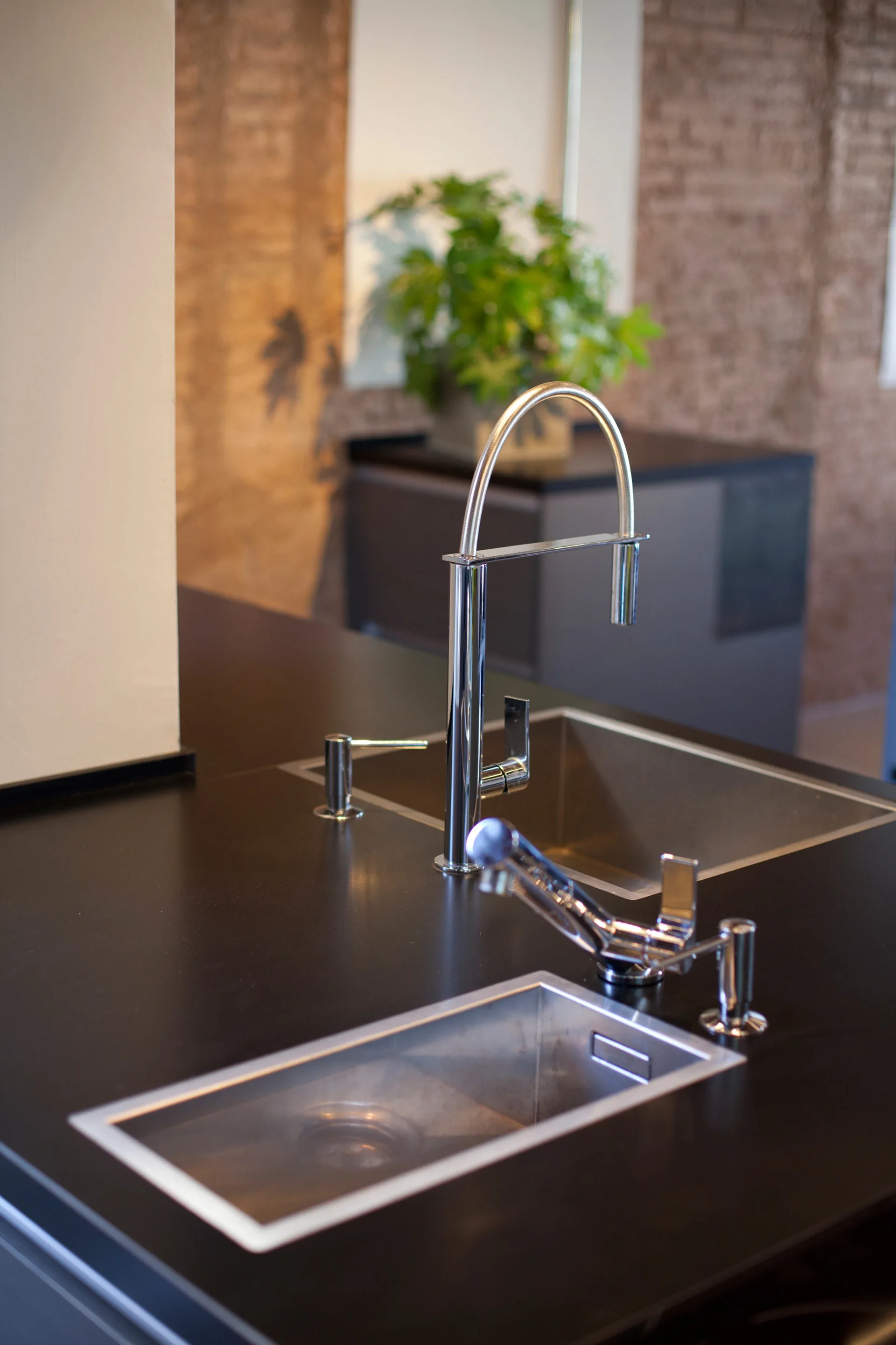 A polished chrome gooseneck faucet and stainless steel undermount sinks are set into a matte black kitchen countertop against an exposed brick wall.