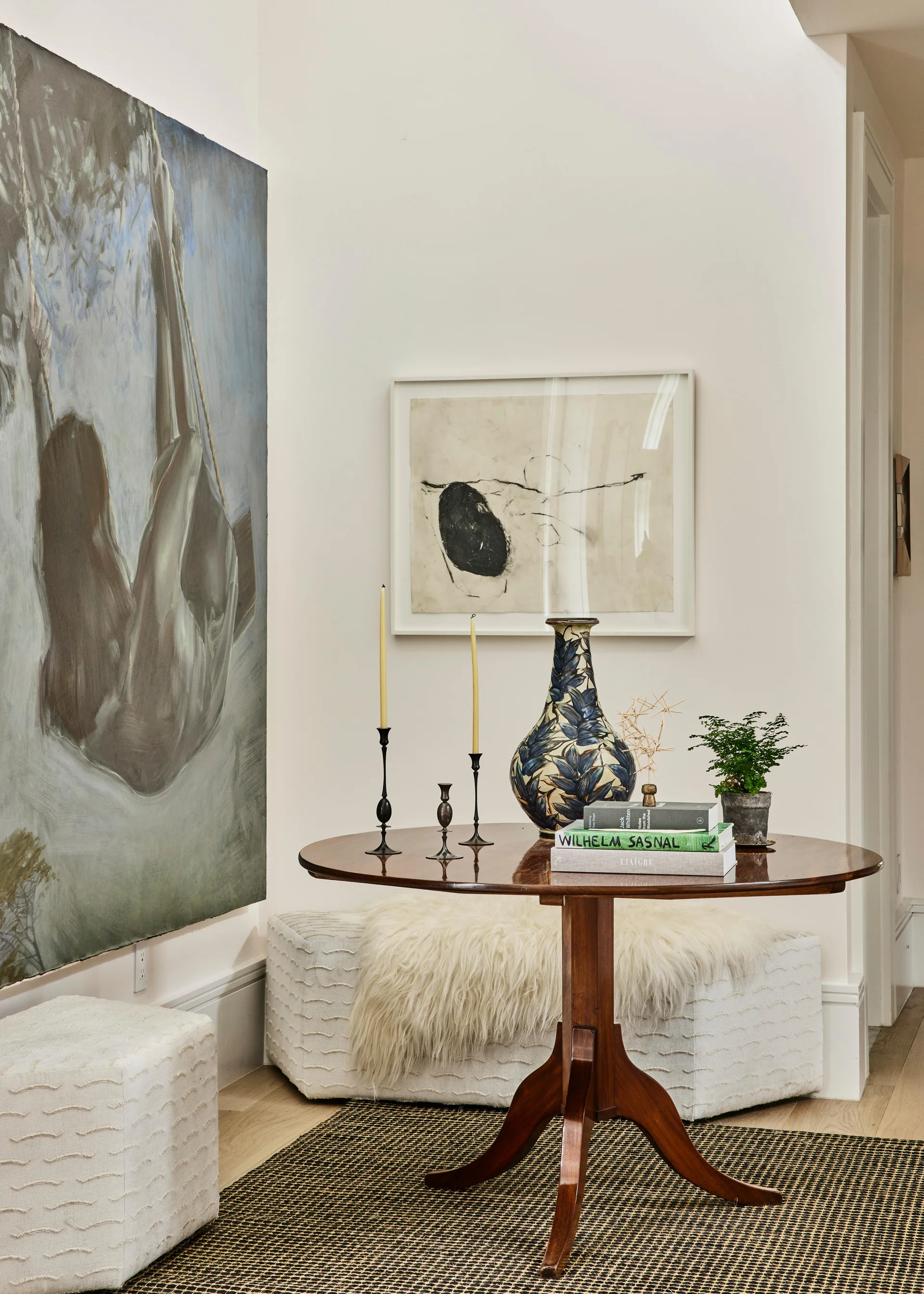 A dark wood pedestal table displaying a blue patterned vase, candlesticks, and books sits on a patterned rug beside textured white ottomans and large wall art.