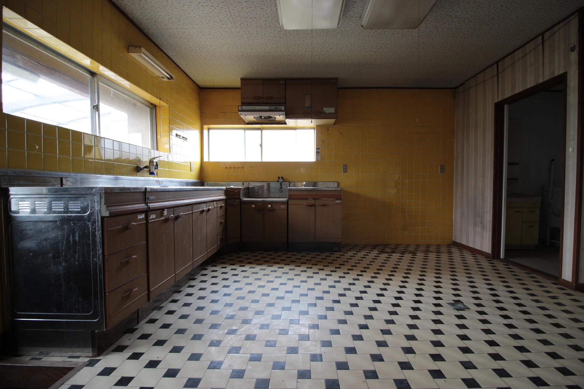A vintage kitchen features glossy yellow tiled walls, wood-grained base cabinets, and a black-and-white checkered vinyl floor.