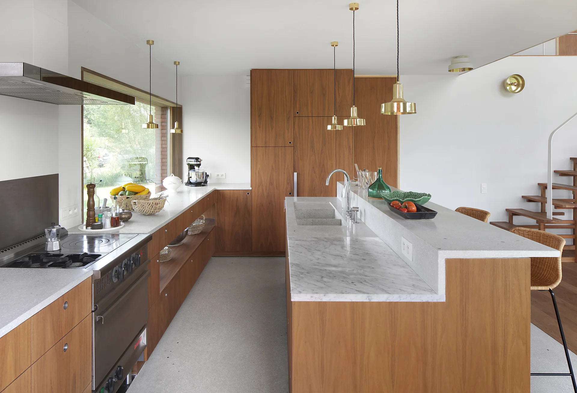 A modern kitchen with flat-panel walnut cabinetry, a tiered island featuring a marble prep surface, brass pendant lights, and a long ribbon window.