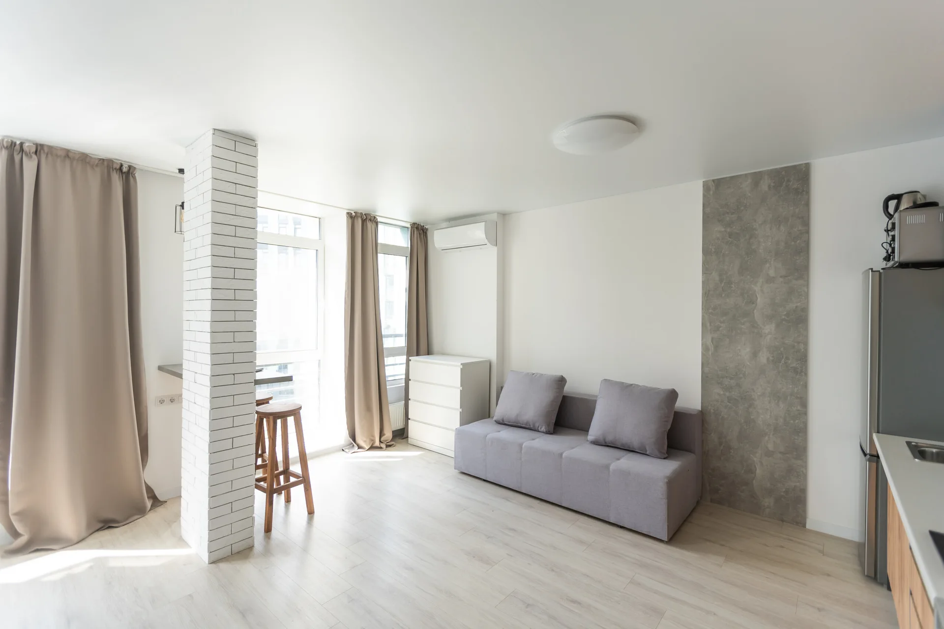 A minimalist living space features a gray fabric sofa, a white brick-clad pillar, and beige curtains beside tall windows.