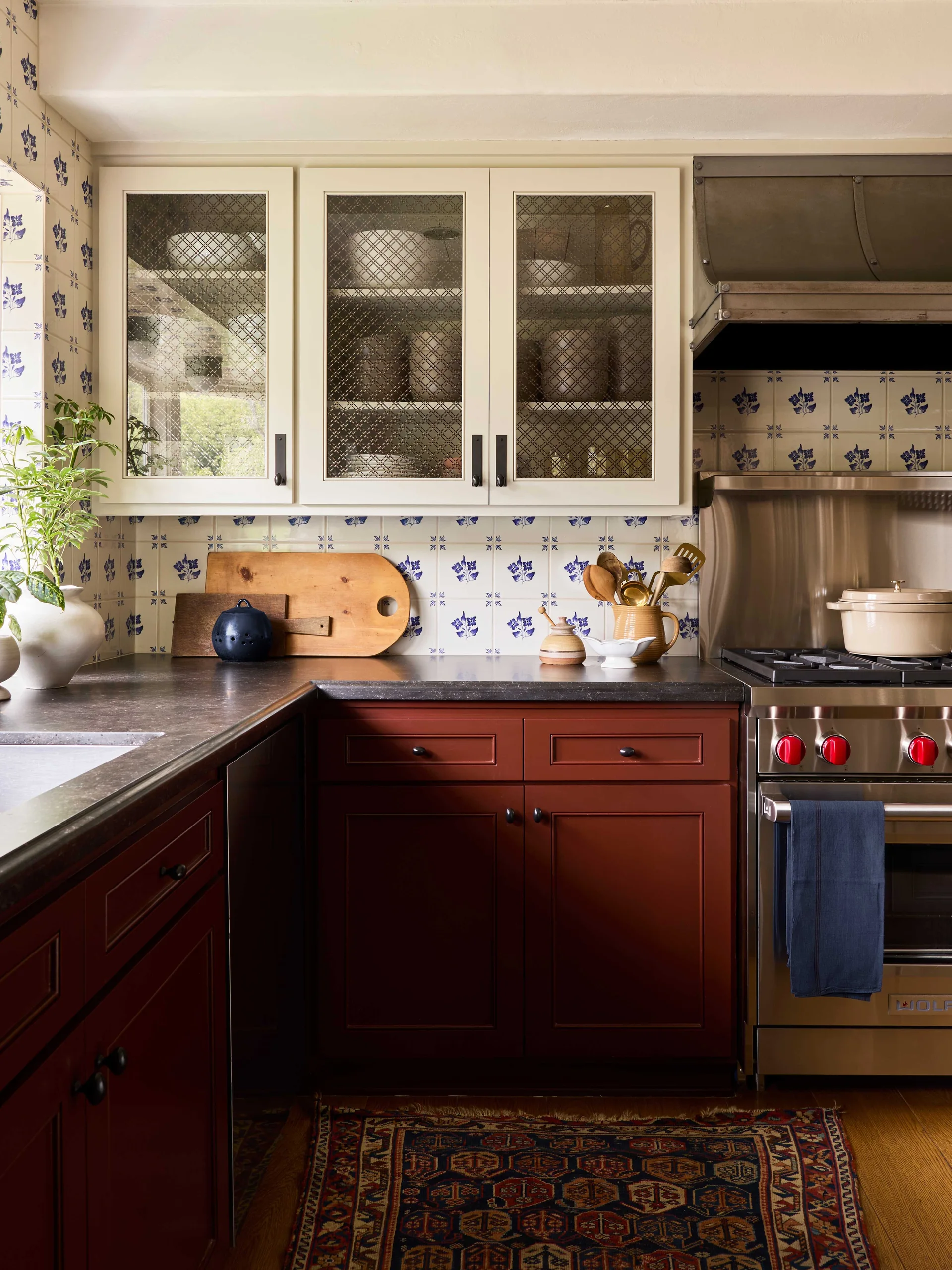 A traditional kitchen featuring deep red base cabinets, cream glass-front upper cabinets with wire grilles, and a blue and white floral tile backsplash.