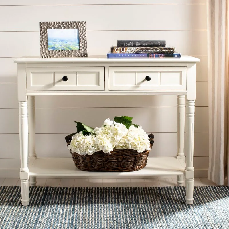 A white wooden console table features a wicker basket of white flowers on its bottom shelf in front of a shiplap wall.