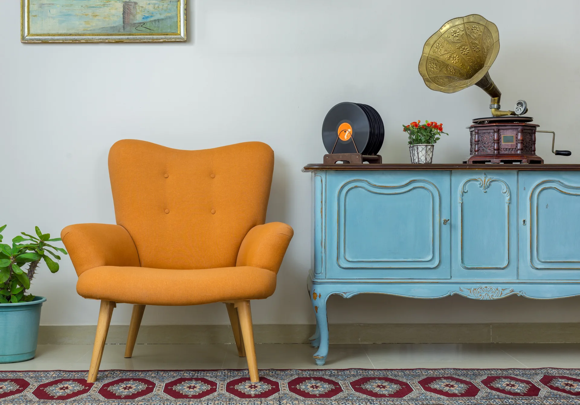 An orange mid-century armchair sits next to a distressed blue wooden sideboard holding a vintage brass gramophone on a patterned geometric rug.