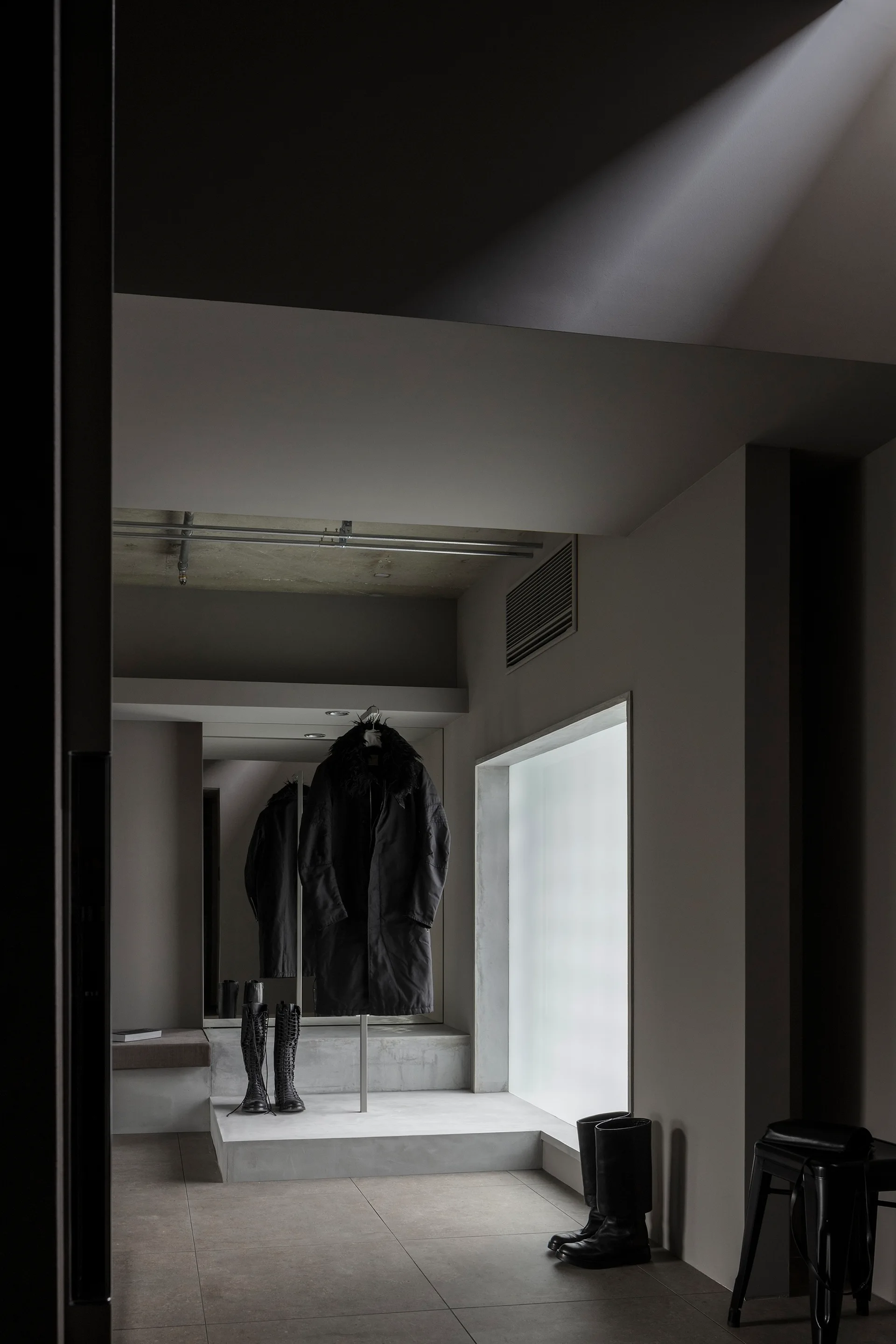 A moody, minimalist dressing room features a raised concrete platform displaying a dark coat, tall boots, a large mirror, and a frosted window.