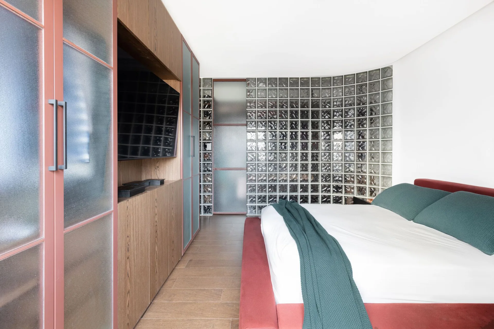 A modern bedroom features a red upholstered bed frame and a wood-toned storage wall with an integrated television set against a curved glass block wall.