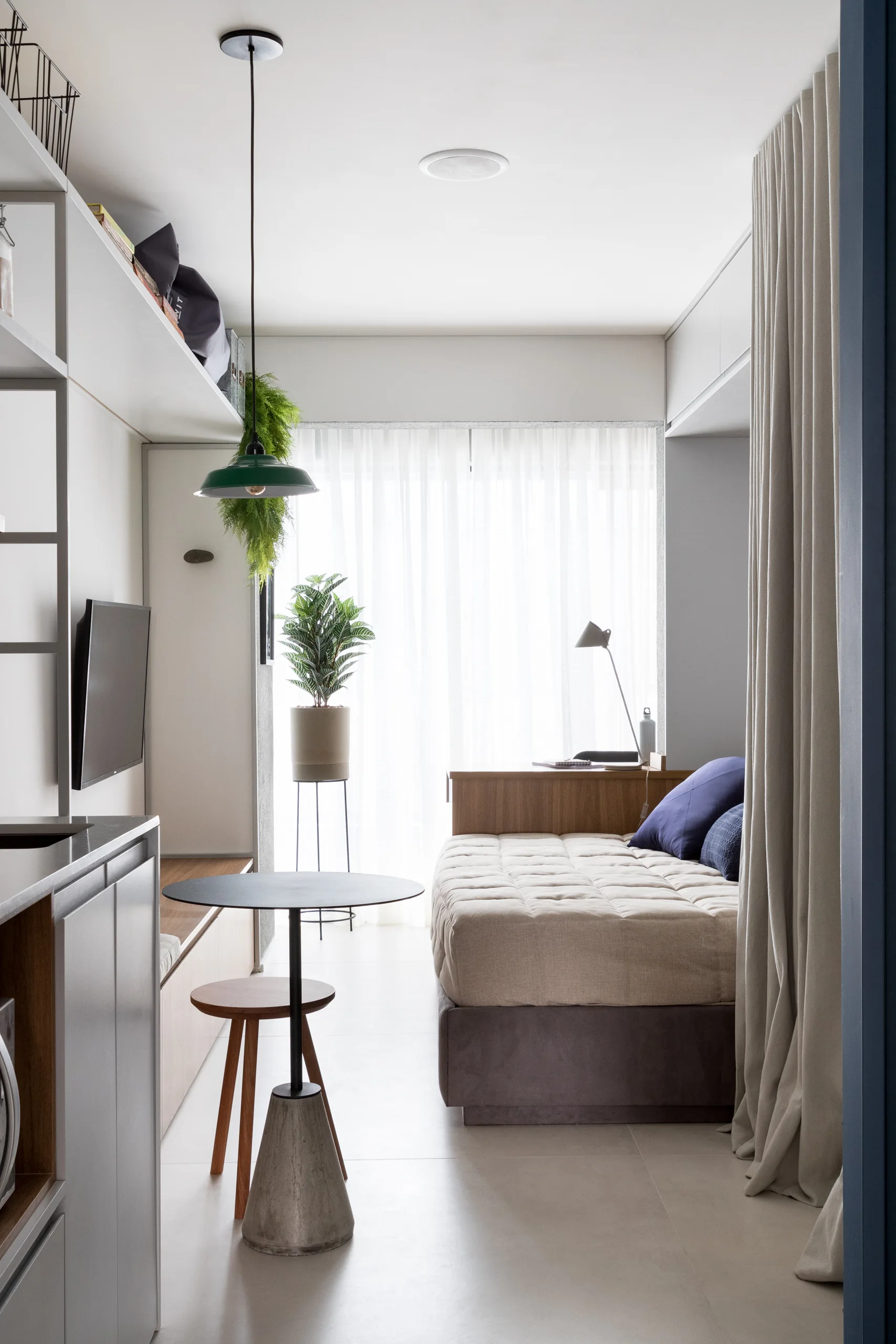 A narrow bedroom featuring a built-in daybed with neutral bedding, floor-to-ceiling curtains, a small bistro table, and extensive white overhead cabinets.