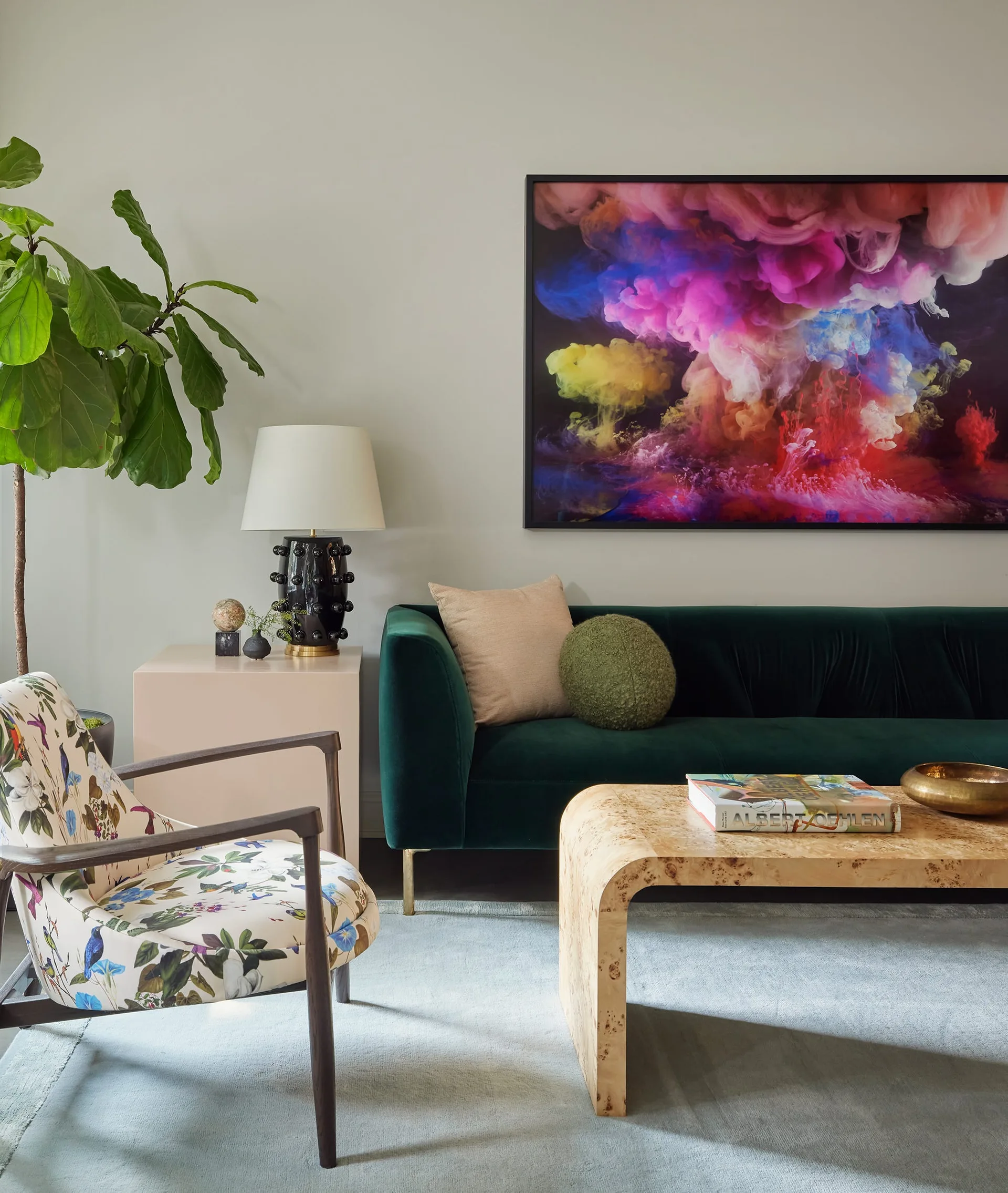 An eclectic living room features a dark green velvet sofa, a floral patterned armchair, and a burl wood coffee table beneath colorful abstract wall art.