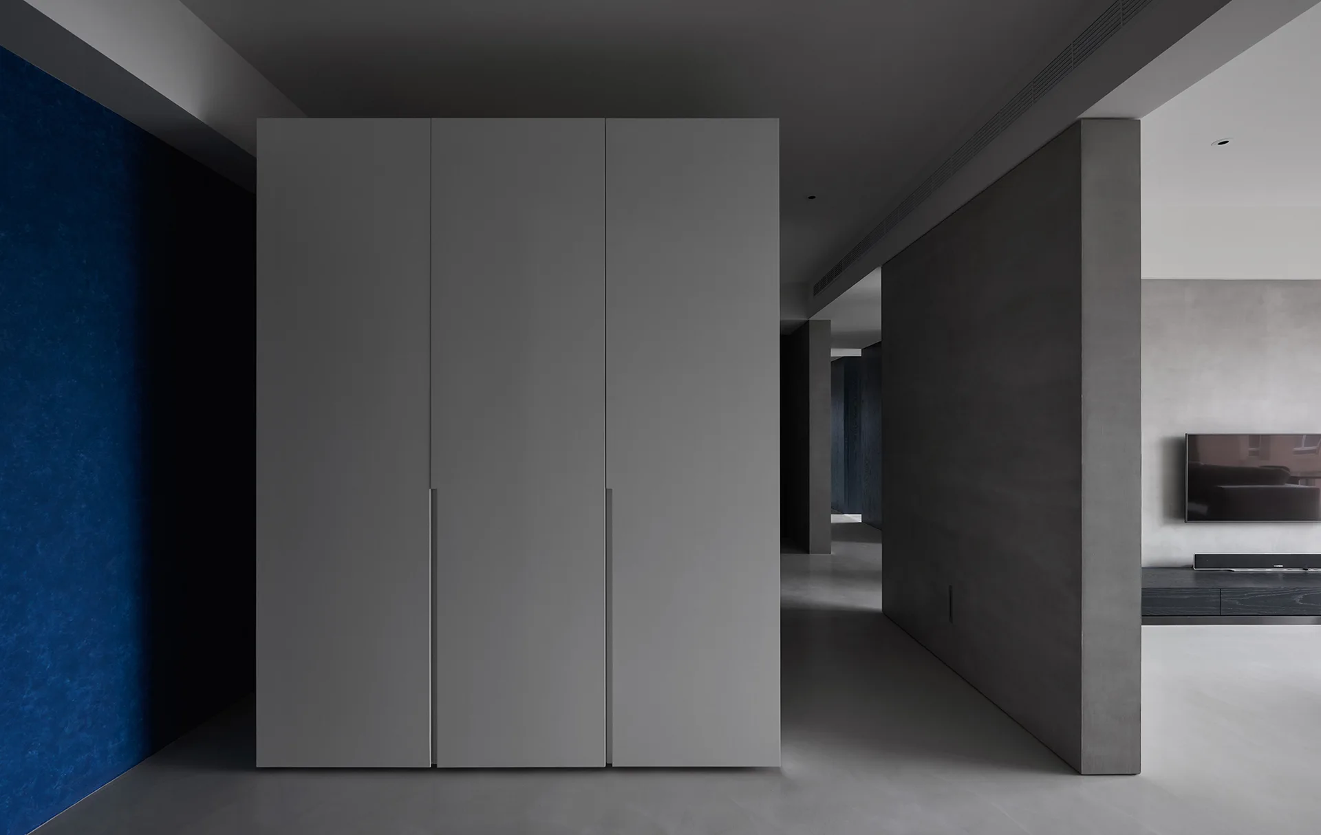 A minimalist gray three-door wardrobe stands near a textured blue accent wall in a modern gray-toned interior with a partitioned hallway.