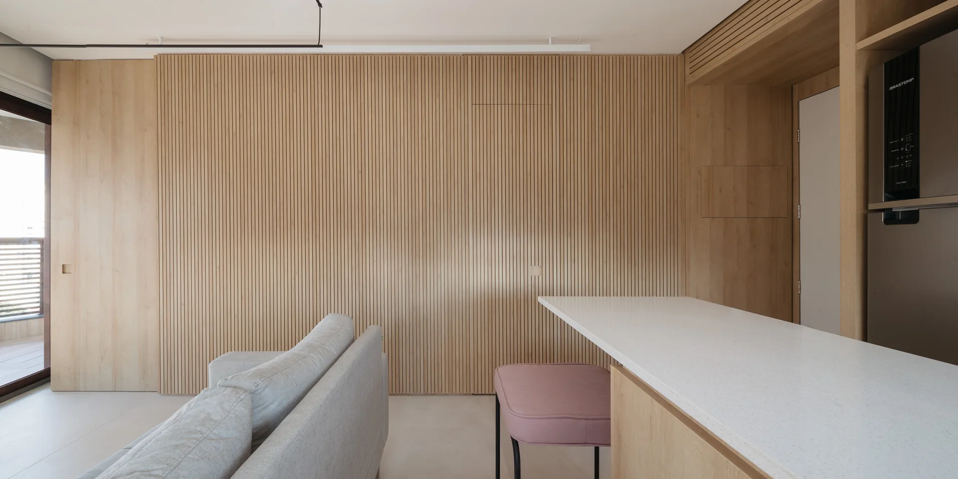 A modern living space features vertical slatted wood wall paneling, a light gray sofa, and a pink stool tucked under a white stone counter.