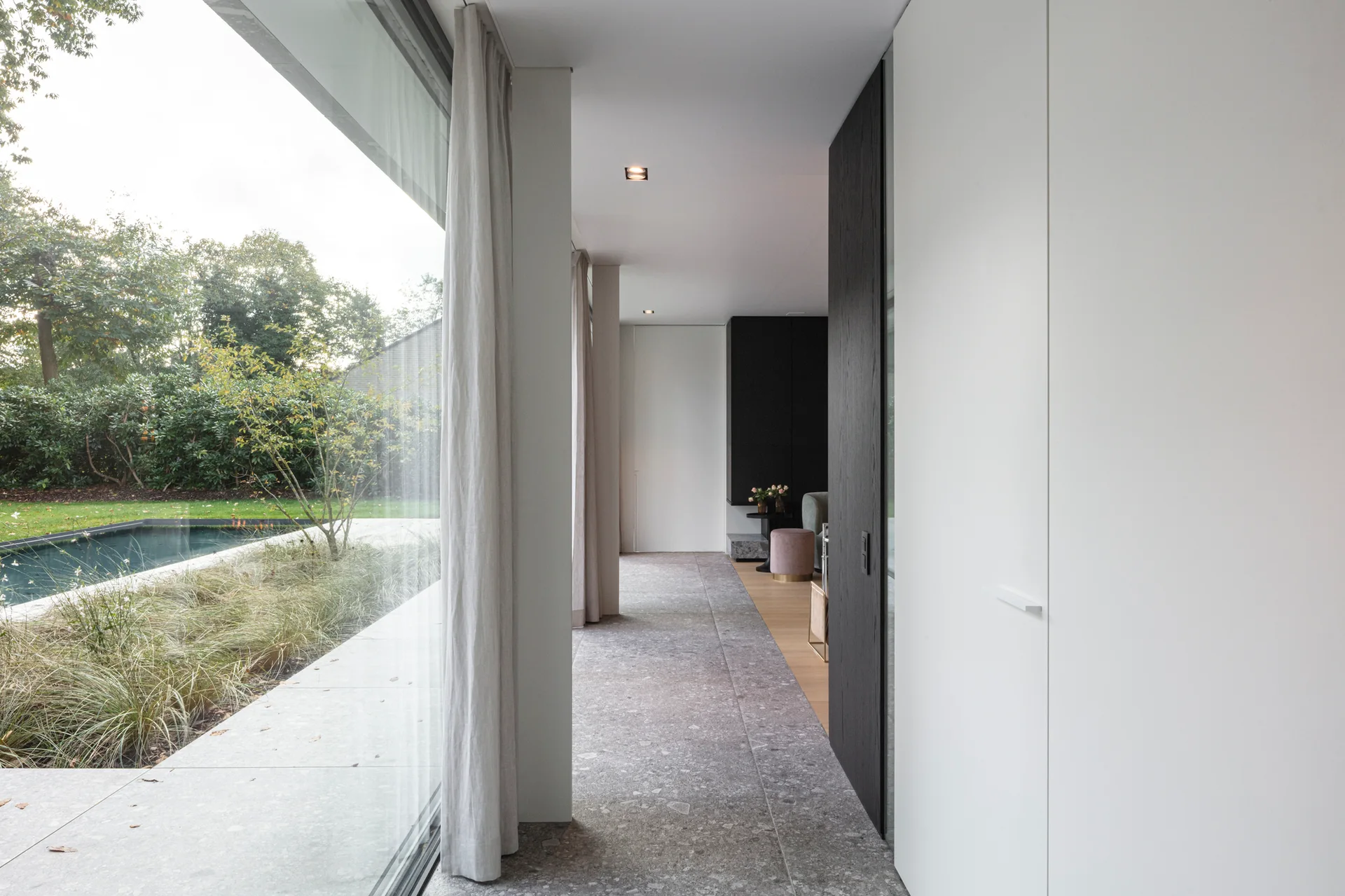 A modern minimalist hallway featuring mottled grey stone flooring, a floor-to-ceiling glass wall with curtains, and flush white storage cabinets.